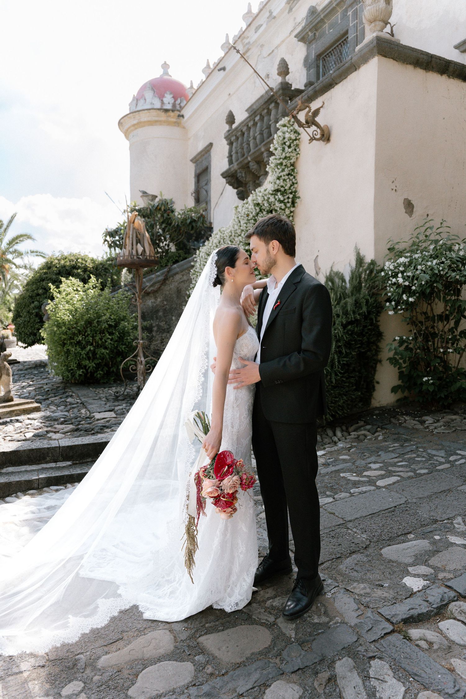 Bride and groom share a romantic moment outside a historic venue, bride holding red bouquet with long flowing veil.