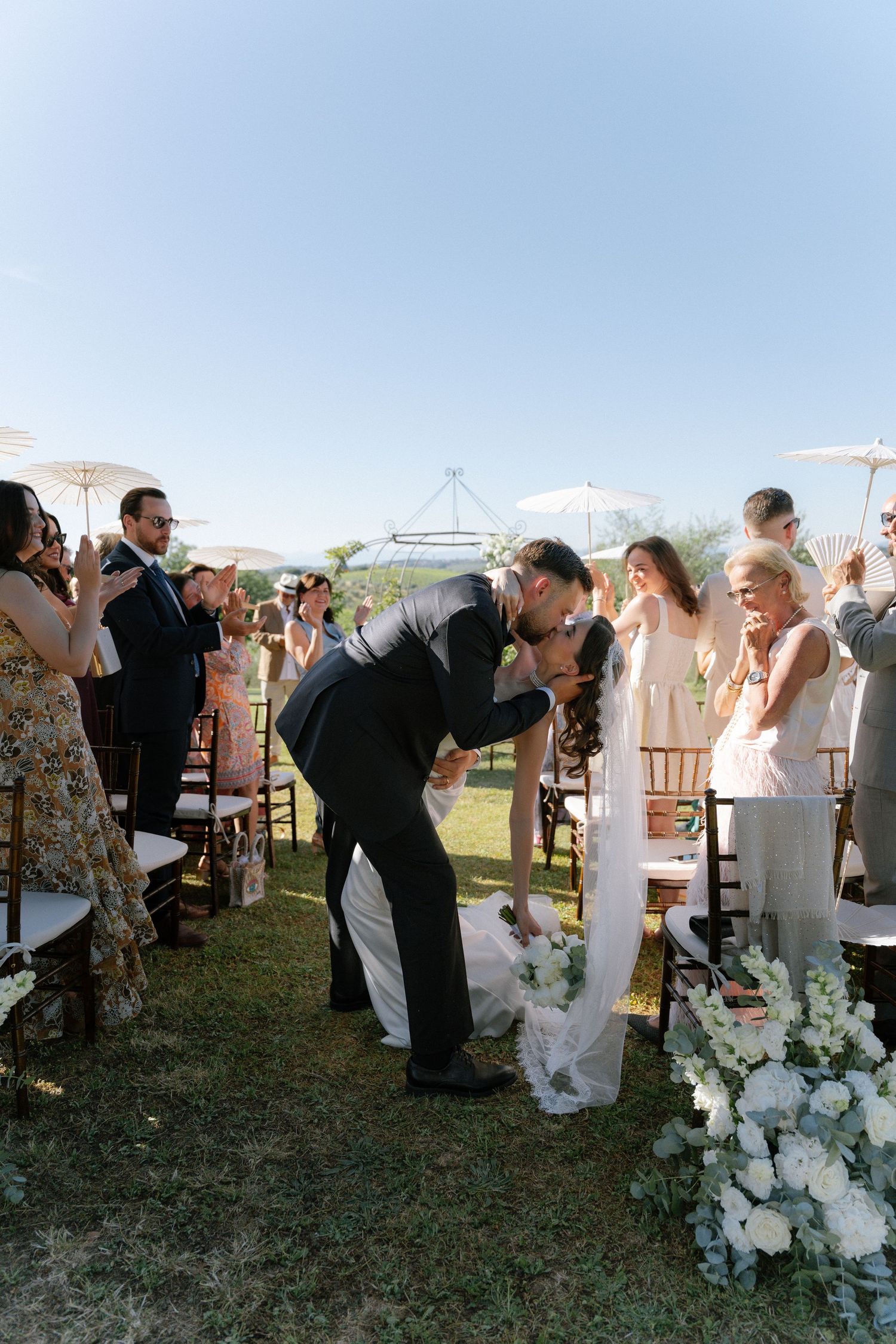 Bride and groom share first kiss at outdoor wedding ceremony with guests applauding on sunny day.