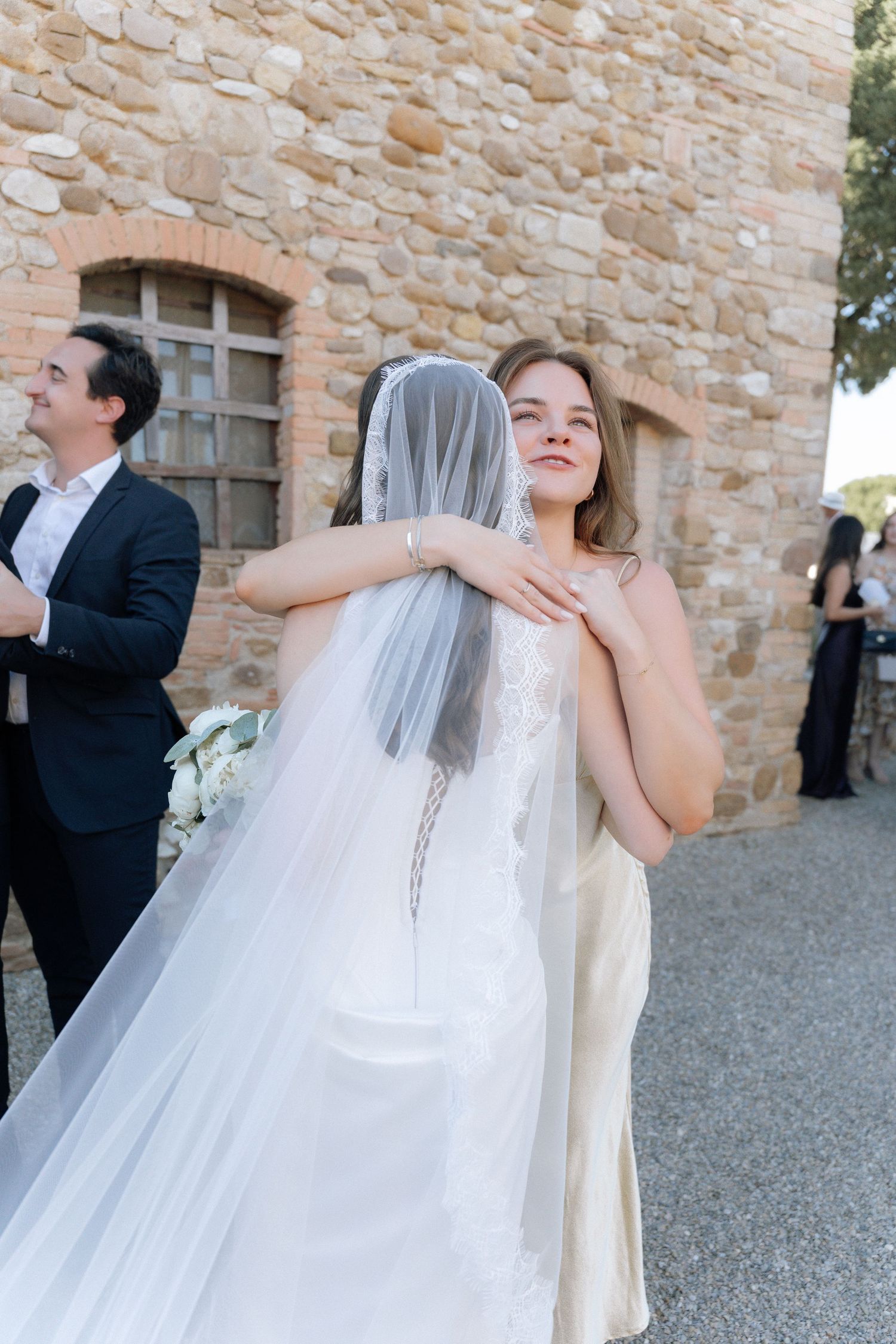 Bride in white gown and veil embraces a guest in champagne dress outside a rustic stone venue.