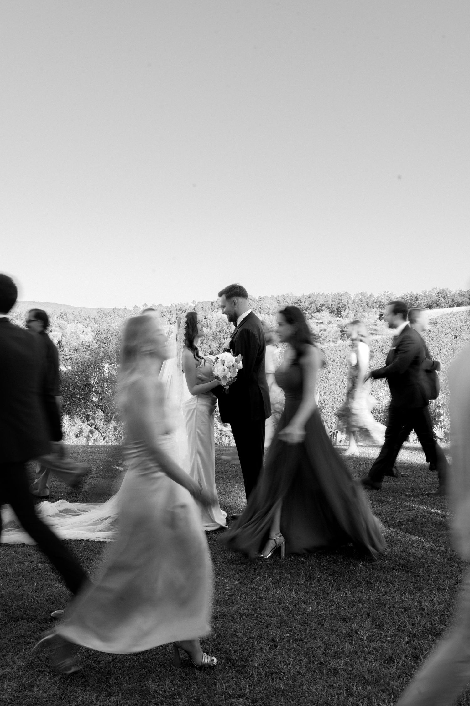 Black and white wedding photo of bride and groom standing still amid blurred moving guests outdoors.
