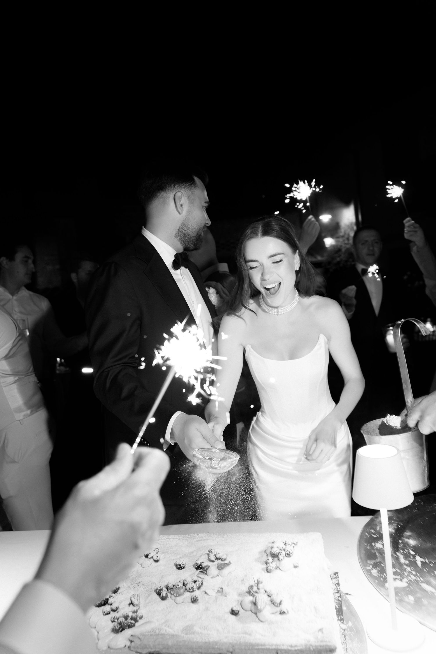 Black and white photo of a bride and groom cutting their wedding cake surrounded by sparklers at a night reception.