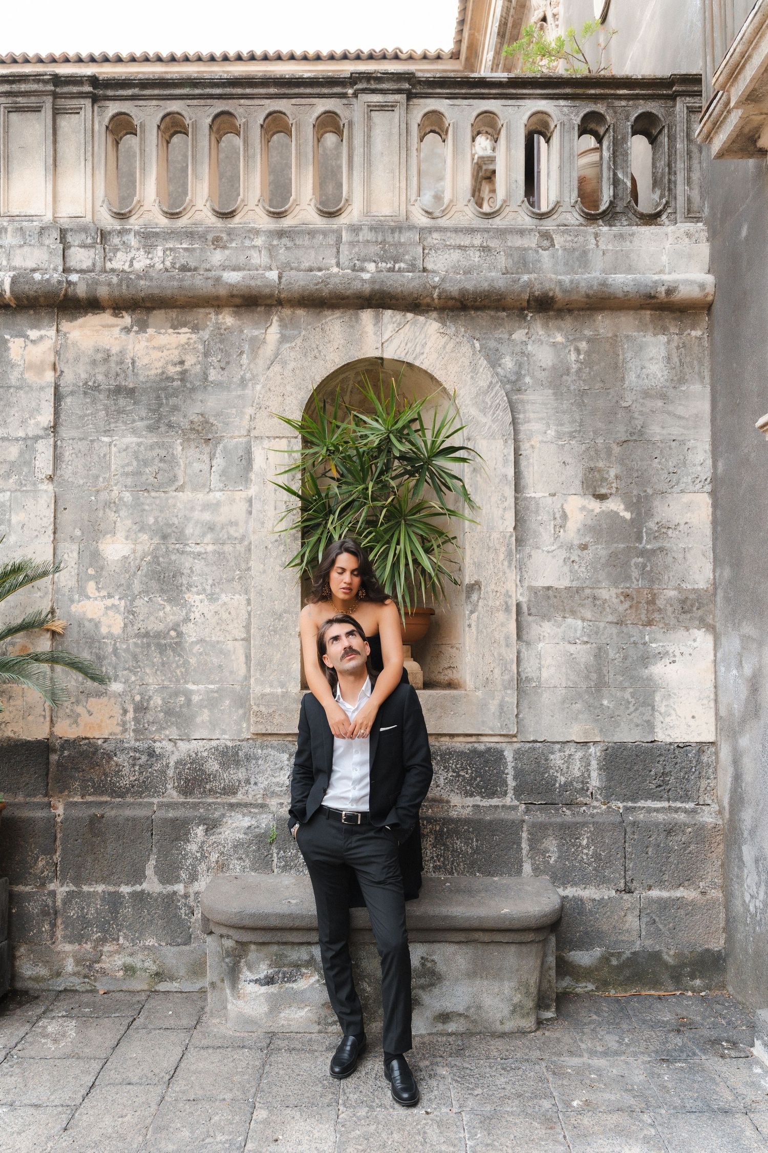 Couple posing against ancient stone wall with tropical plants, man in black suit, woman in white top, outdoor setting.
