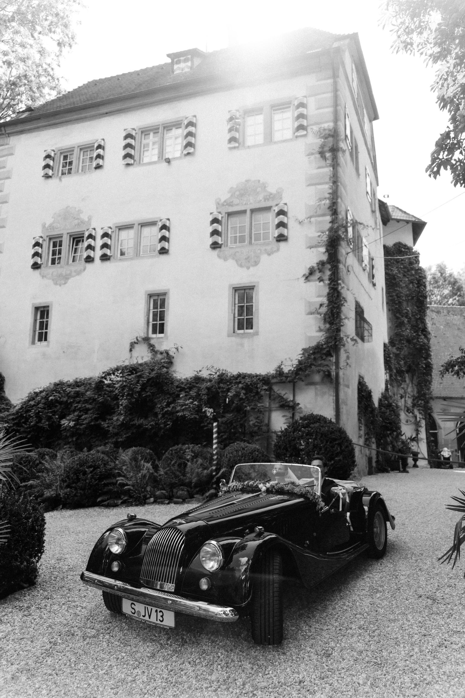 Black and white photo of a vintage convertible car decorated for a wedding parked on a gravel drive in front of a historic manor house.