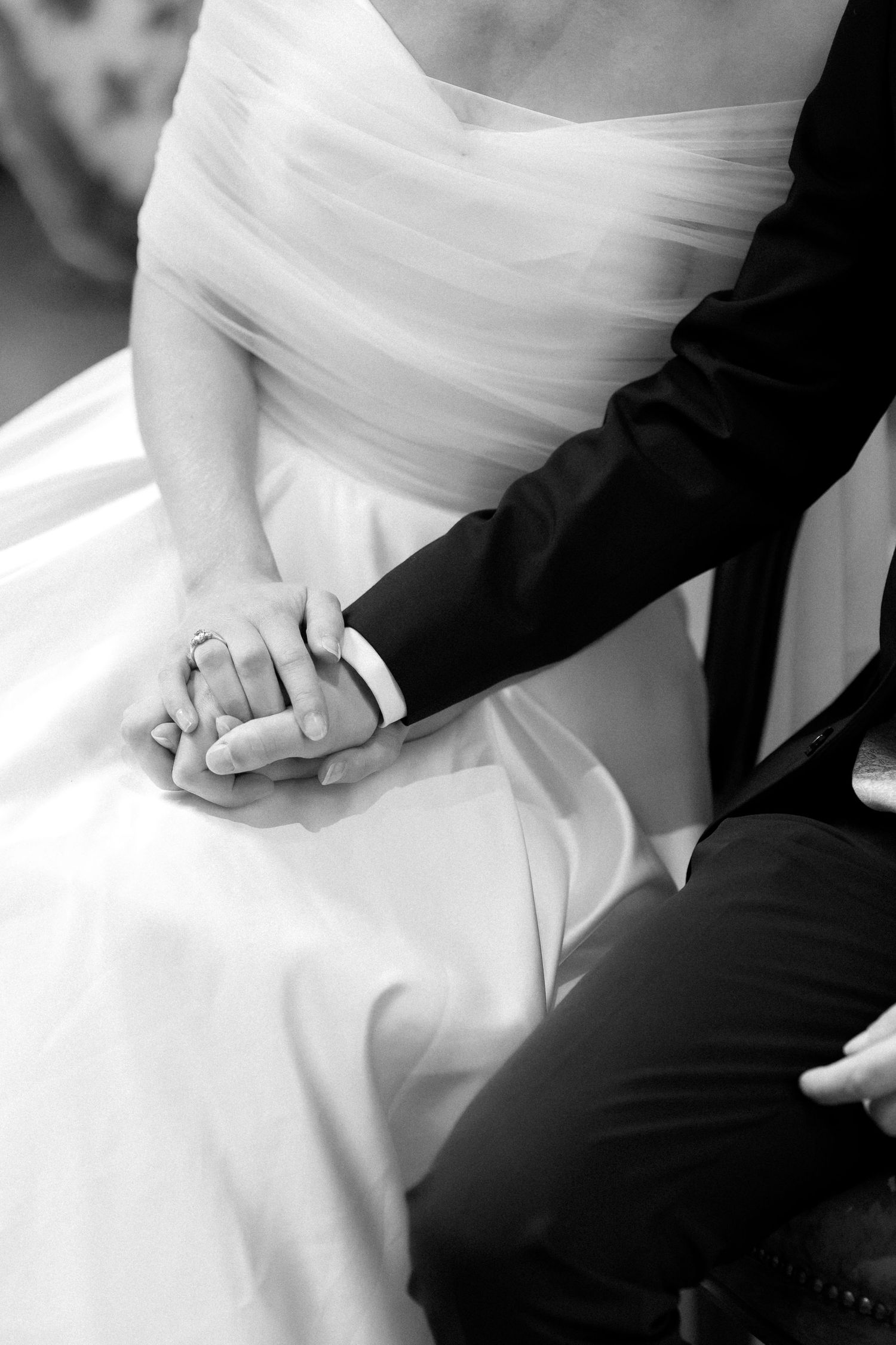 Black and white photo of a bride in a tulle wedding gown holding hands with a groom in a dark suit, her ring visible.