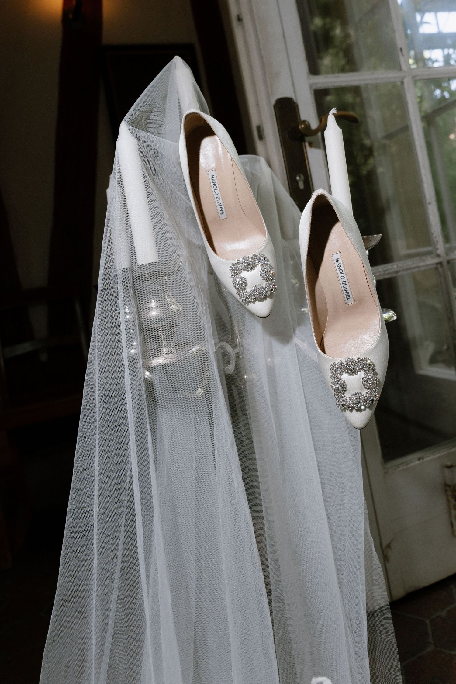 White satin bridal heels with jeweled crystal buckles hang beside a flowing tulle wedding veil and candle holder by a glass door.