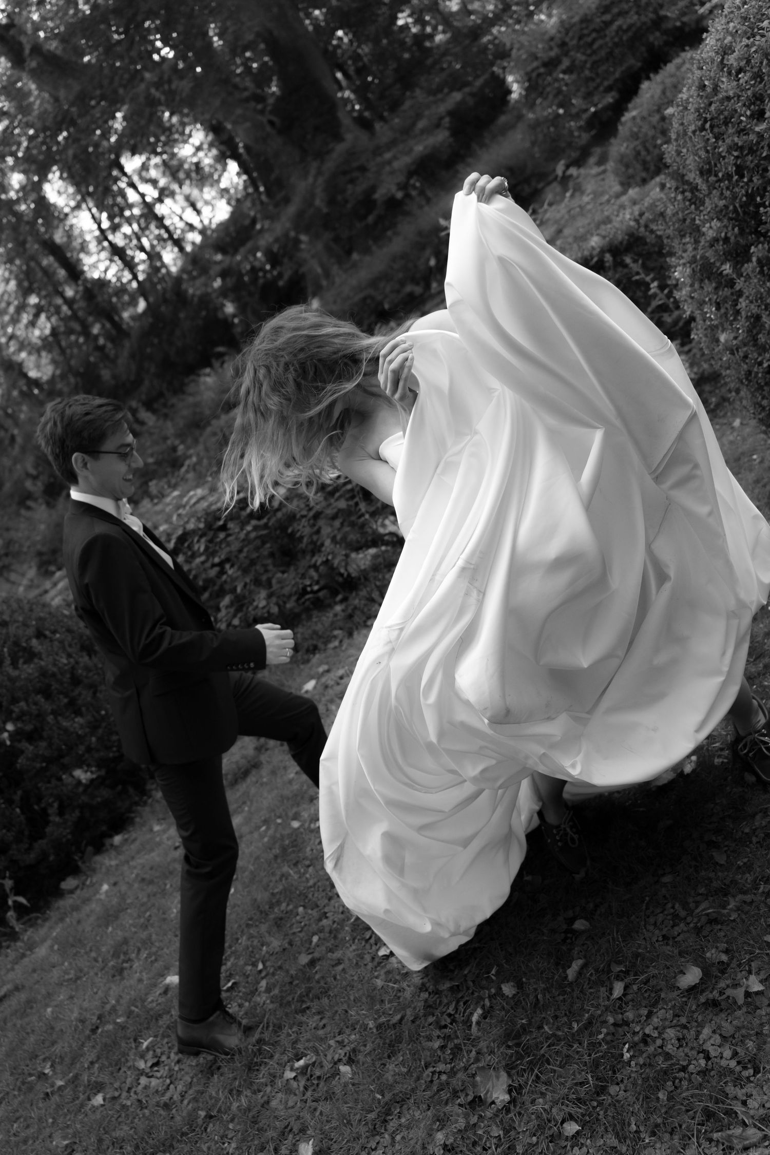 Black and white photo of a bride in a flowing white gown twirling on a grassy hillside beside a groom in a dark suit.