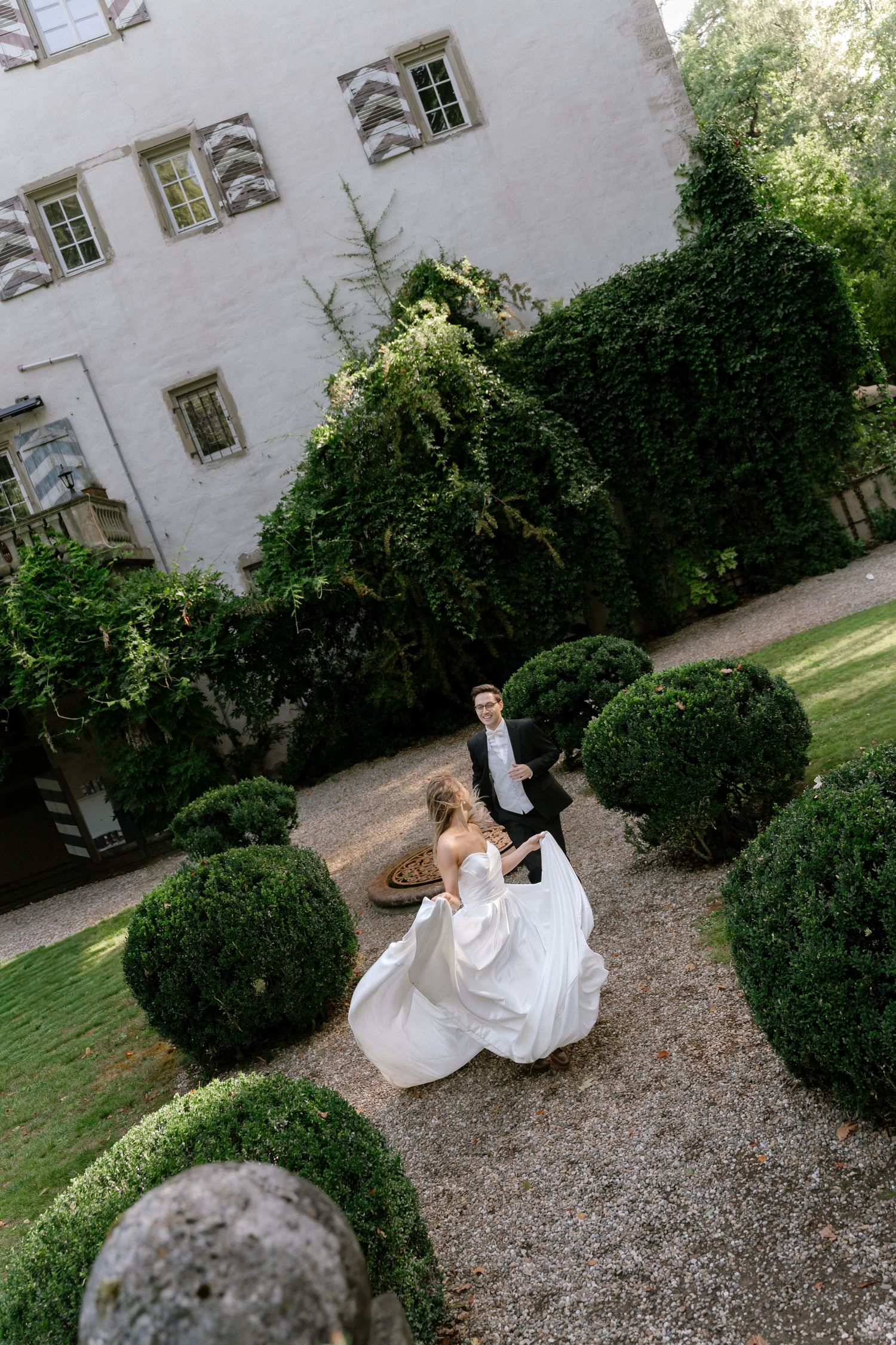 Bride in a flowing white gown twirls on a gravel path past trimmed round topiaries as the groom watches near an ivy-covered manor.