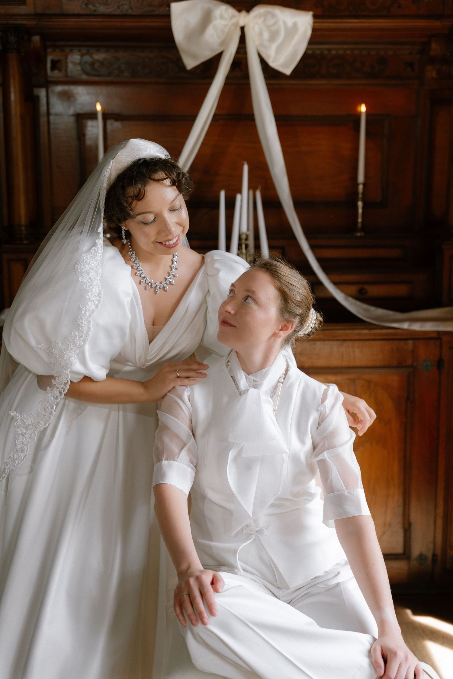 Two brides in white gowns share a tender moment in an elegant wood-paneled room with candles and ribbon decor.