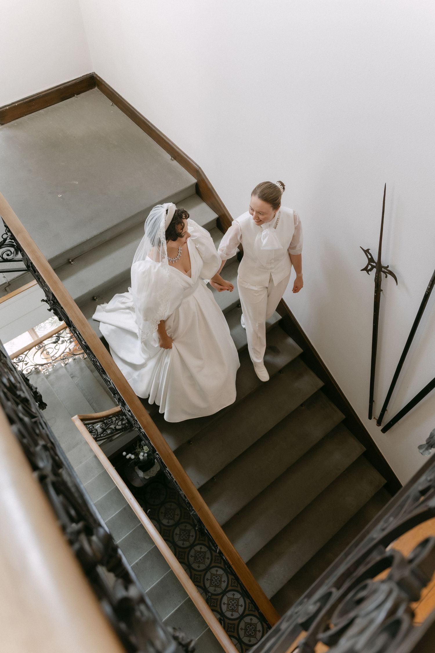 Two brides in white wedding attire holding hands while ascending an elegant staircase with ornate iron railings.