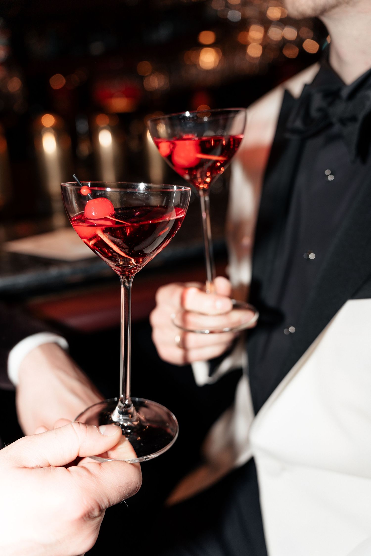 Two hands holding elegant martini glasses with red cocktails against a candlelit bar setting.