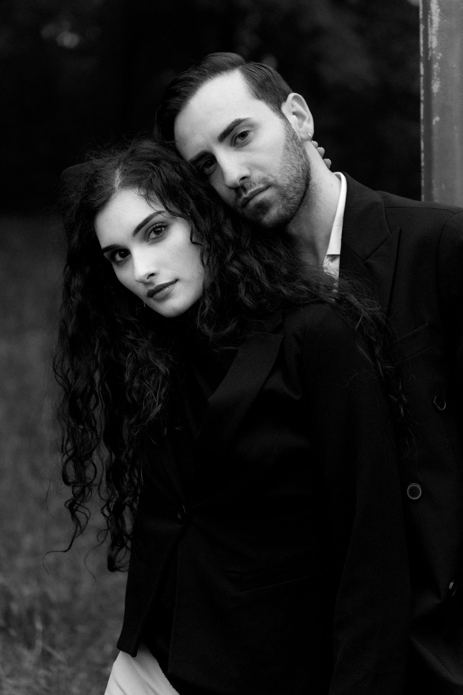 A dramatic black and white portrait of a couple in dark formal attire, with long curly hair and intense expressions.