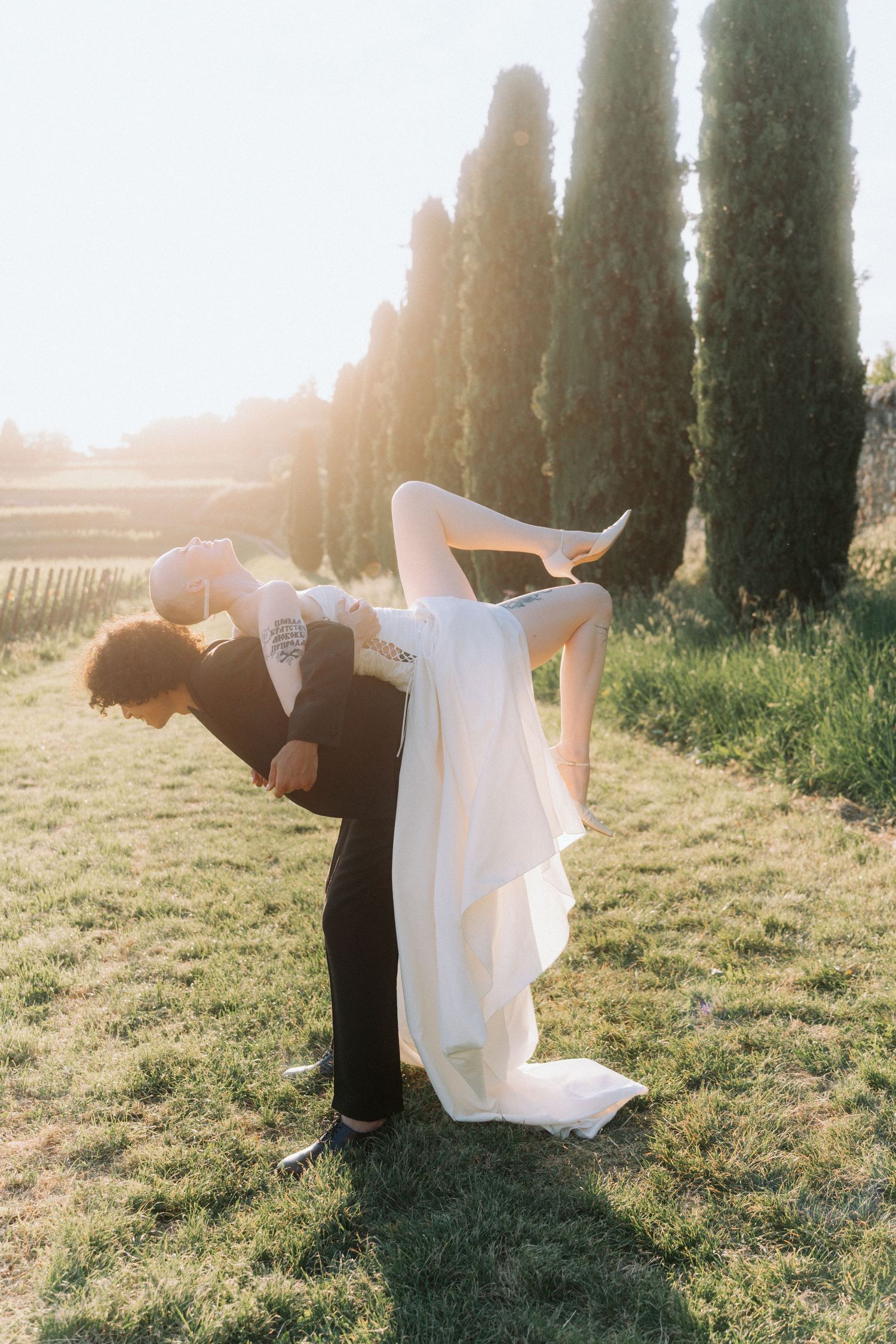 A couple poses dramatically in a Tuscan garden with cypress trees and golden sunlight during their wedding photoshoot.