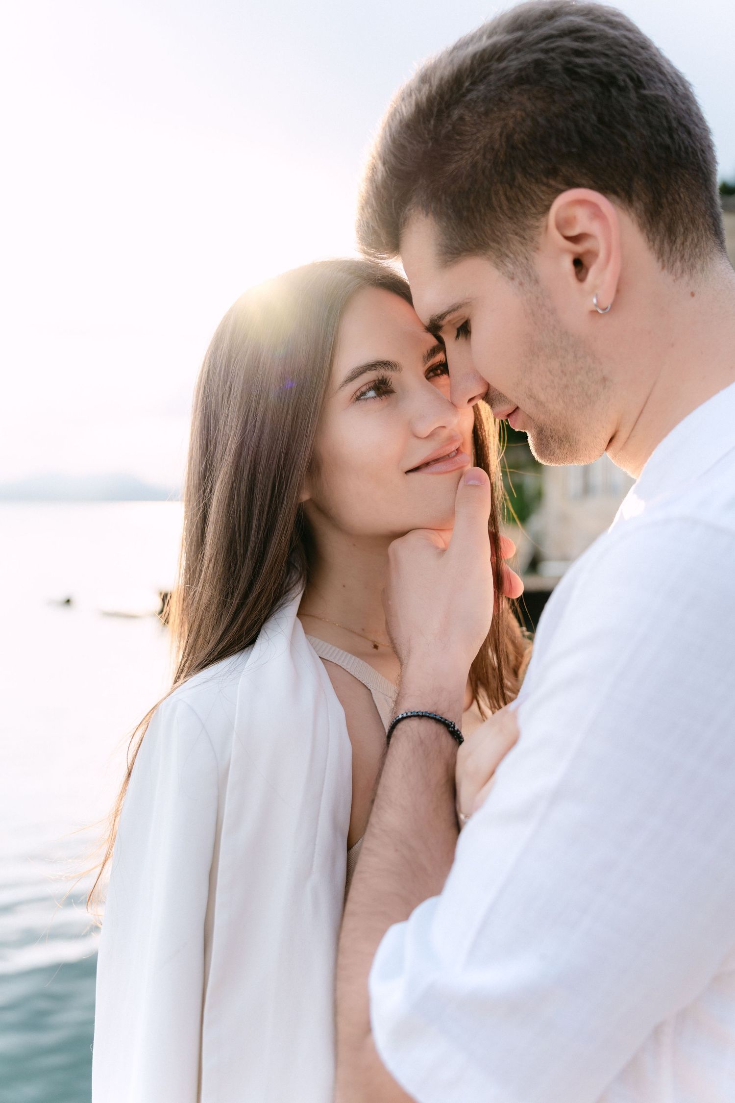 A romantic couple in white clothing share an intimate moment by the water at sunset.
