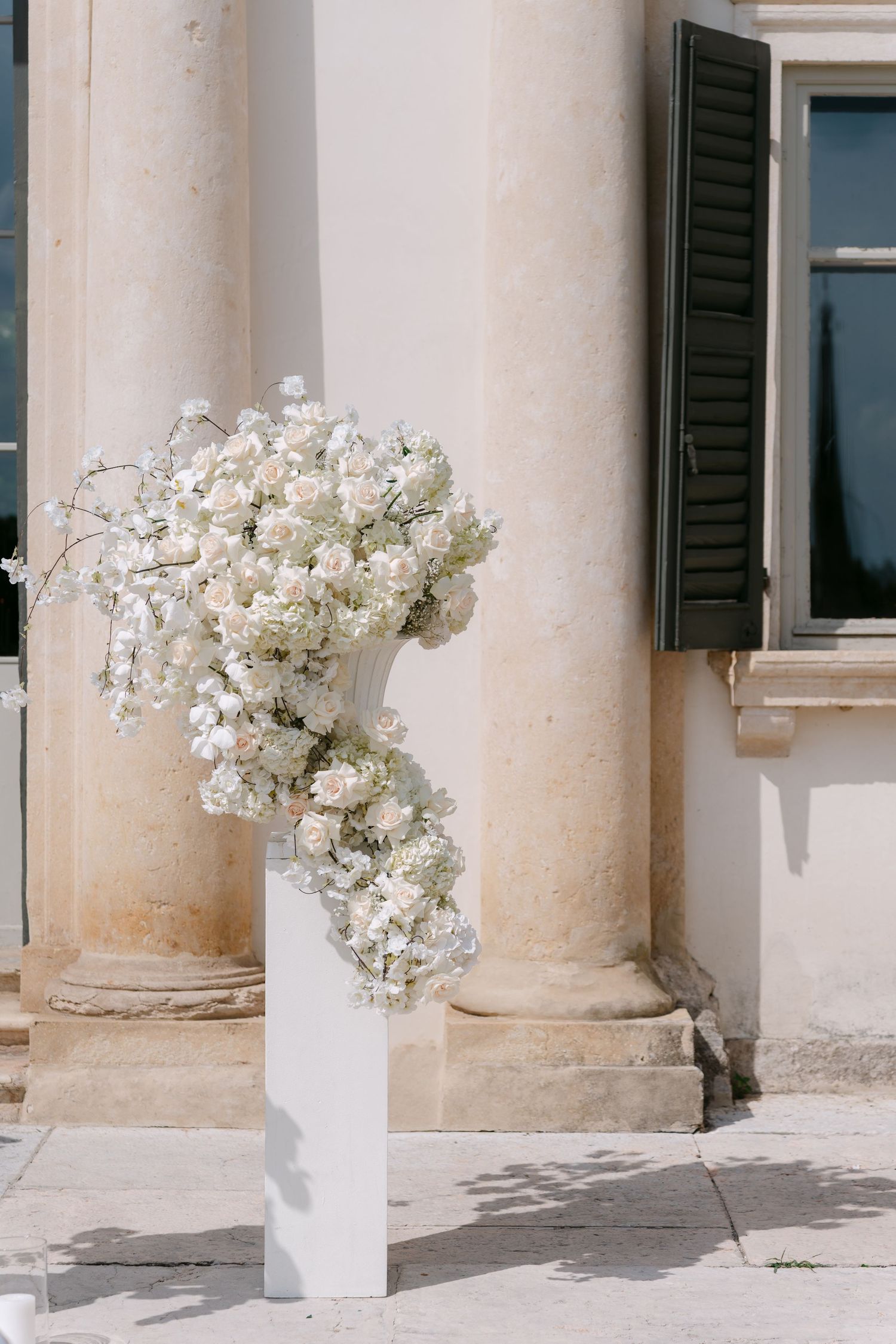 White hydrangea flowers in vases line a row of elegant stone columns along a Mediterranean-style building exterior. Villa Mosconi Bertani