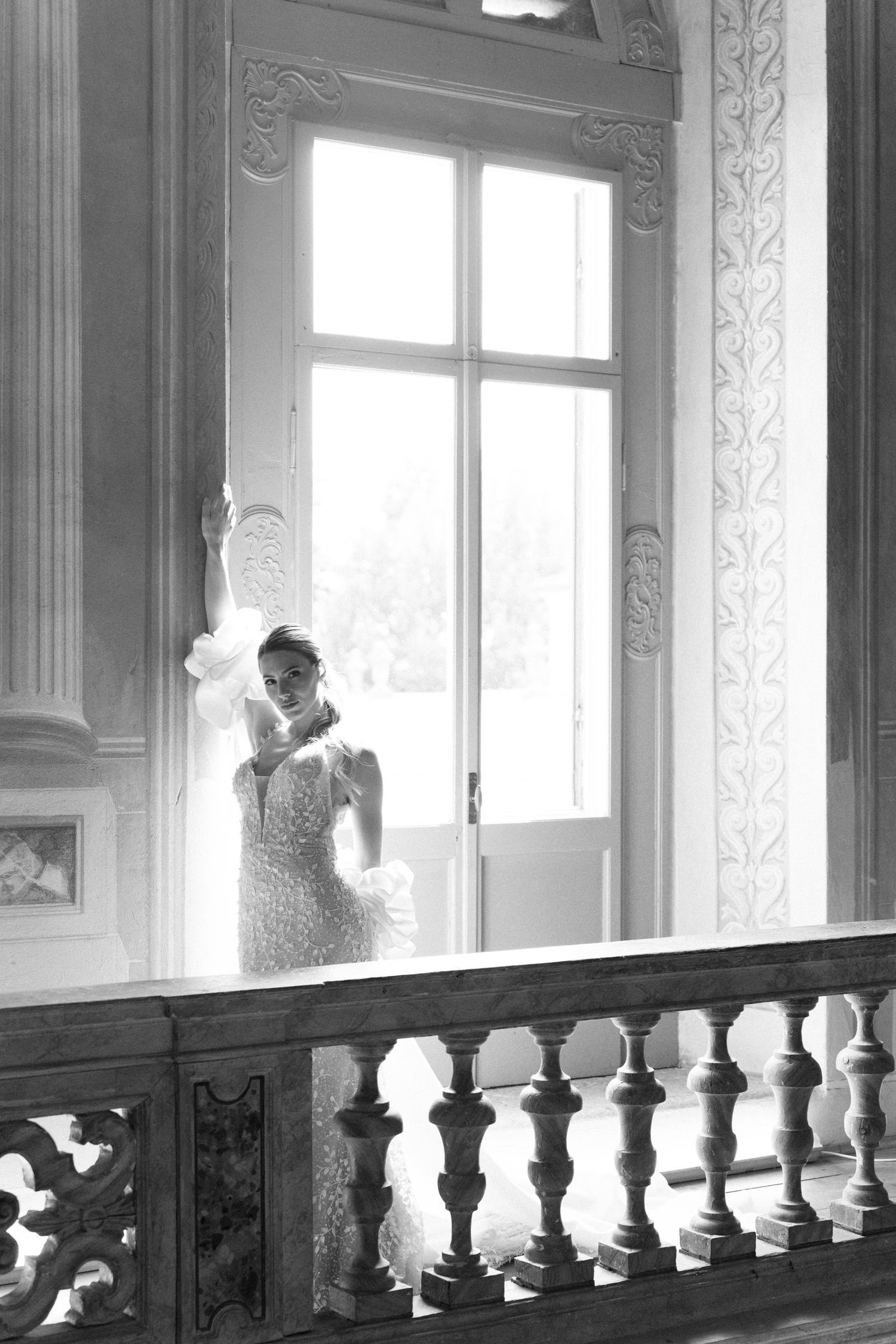 A black and white photograph of an elegant figure in a lace dress standing by an ornate window with decorative balustrade. Wedding Verona