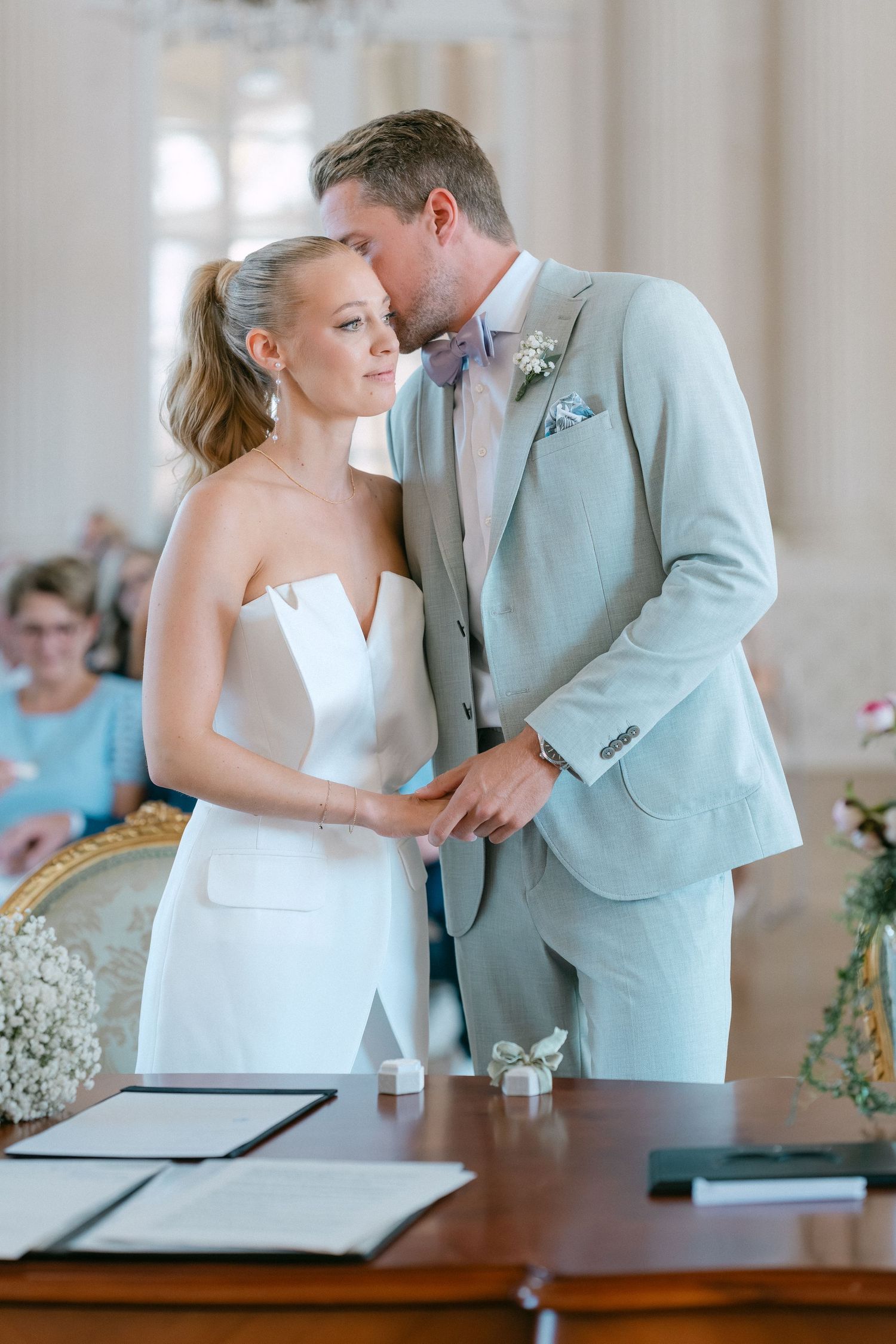 A tender moment between newlyweds at a wedding ceremony signing table with white and light blue attire. Schloss Solitude
