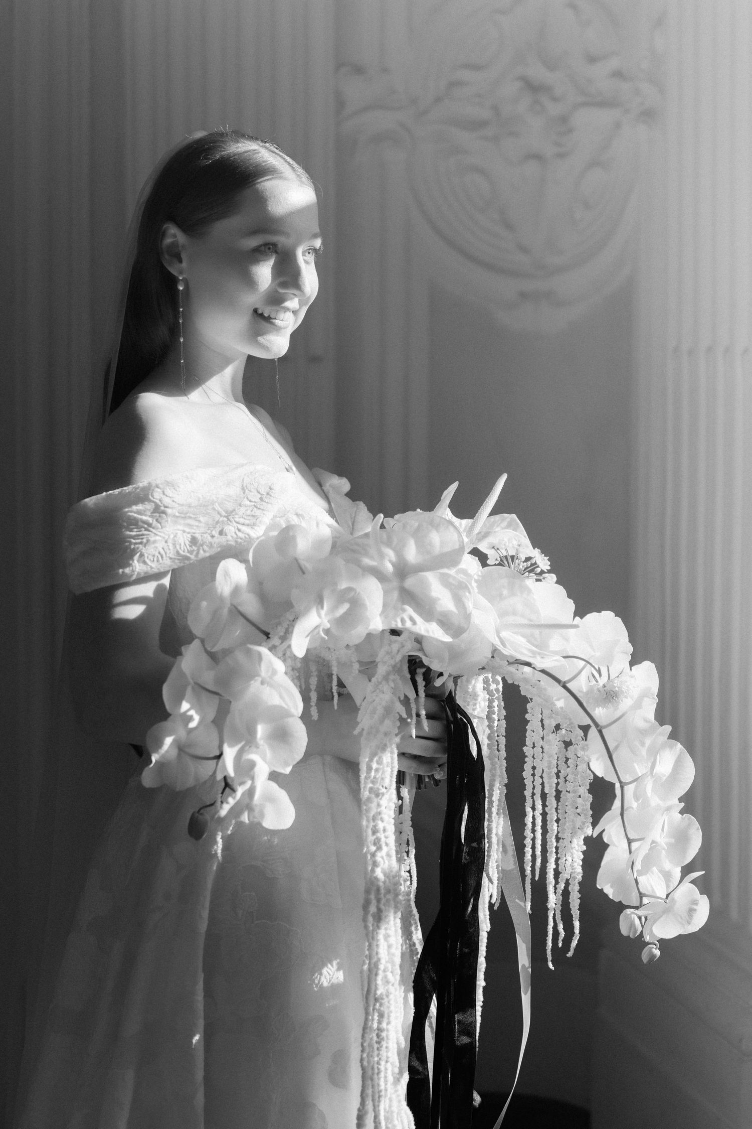 Black and white photo of a smiling bride in an off-shoulder gown holding a cascading orchid bouquet with ribbon streamers.