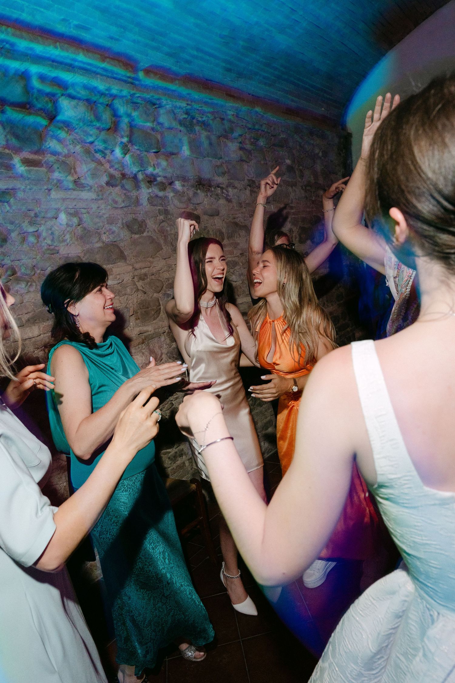 Women in elegant dresses dancing joyfully at a party in a stone-walled venue with colorful lighting.