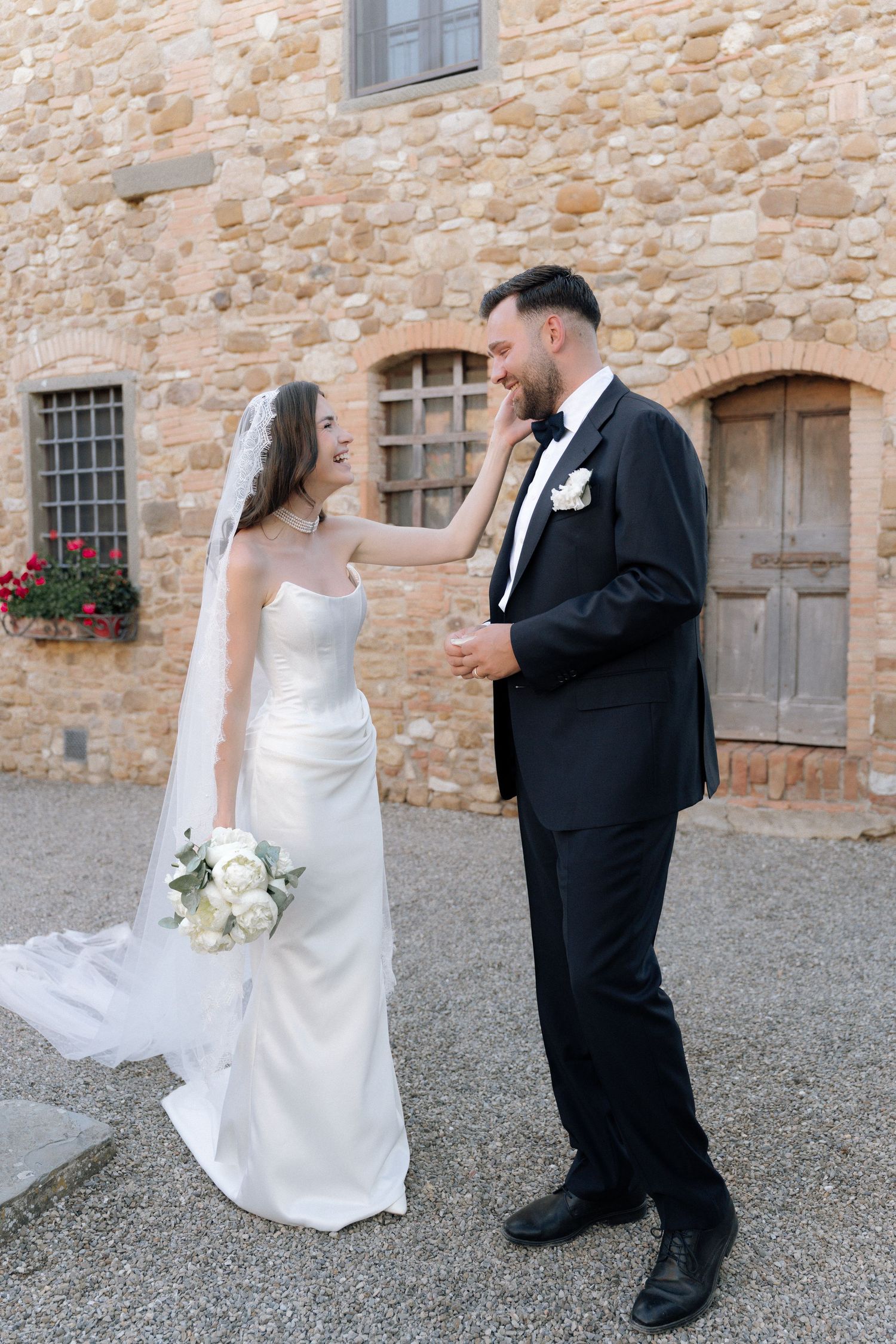 Bride and groom share a romantic moment outside a rustic stone building, bride holding white bouquet in elegant gown.