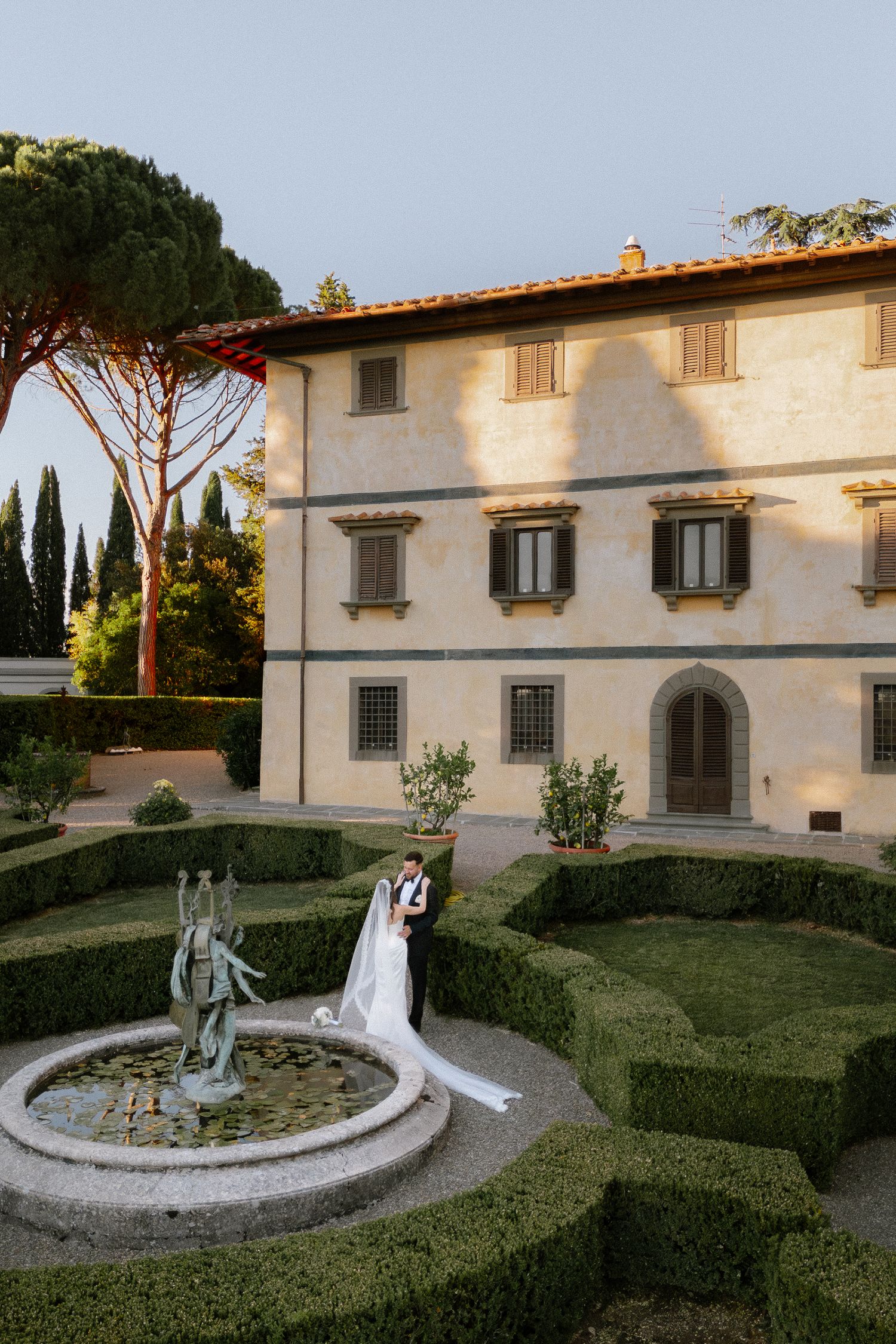 Bride and groom embrace in a formal Italian villa garden with sculpted hedges and a classical fountain at golden hour.
