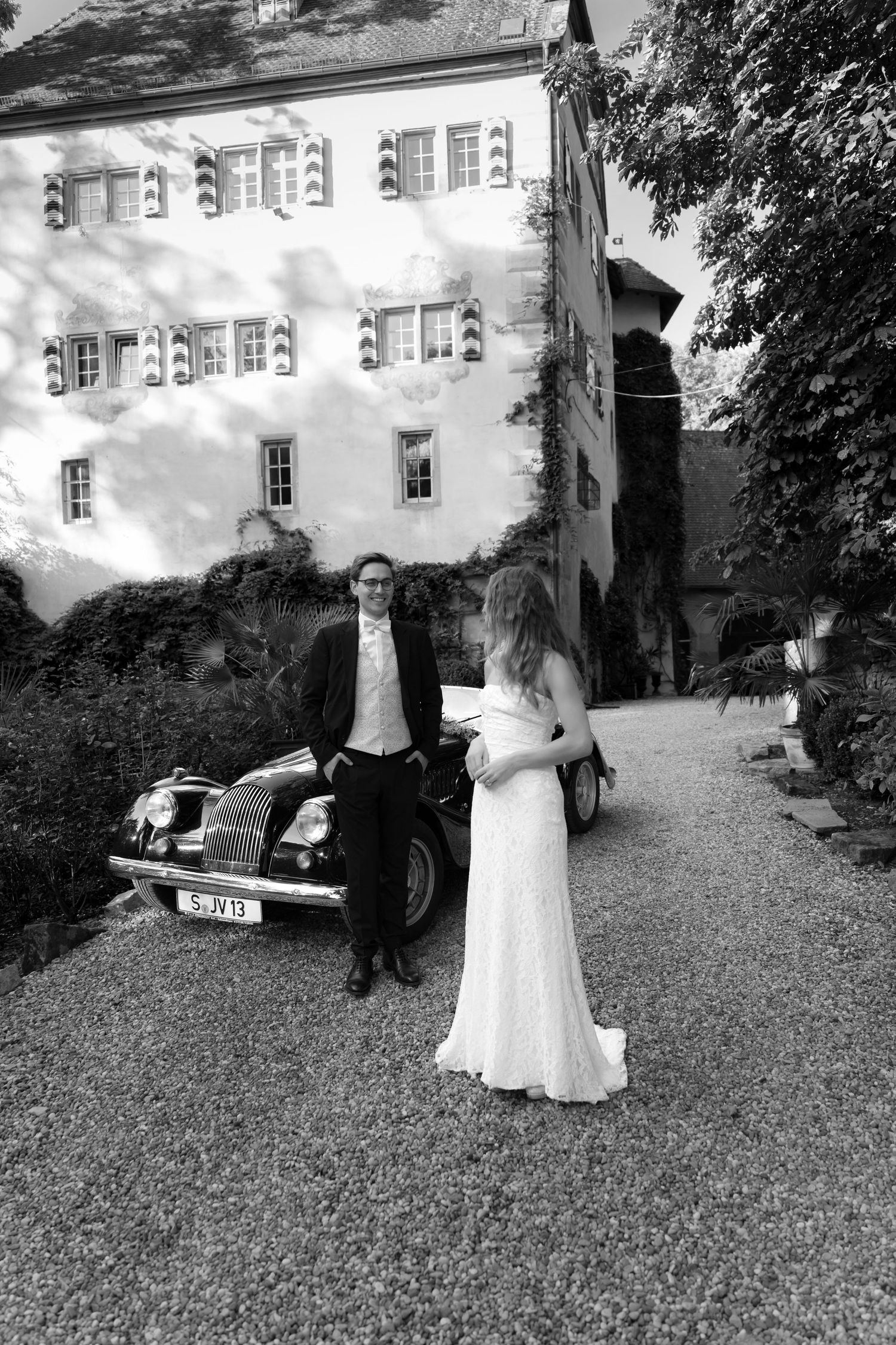 Black and white wedding photo of a bride in a gown and a groom in a suit standing beside a vintage car in front of a historic castle.