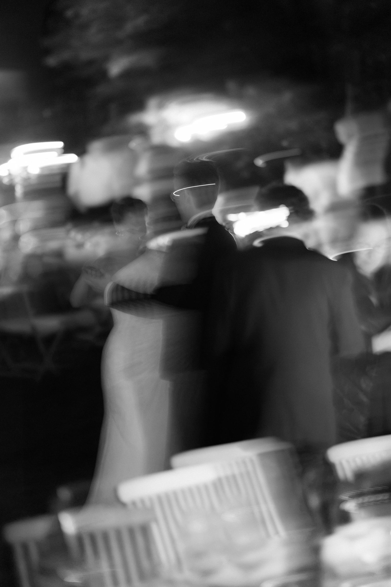 Blurred black and white photo of a couple in formal attire embracing on a dance floor with white chairs in the foreground.