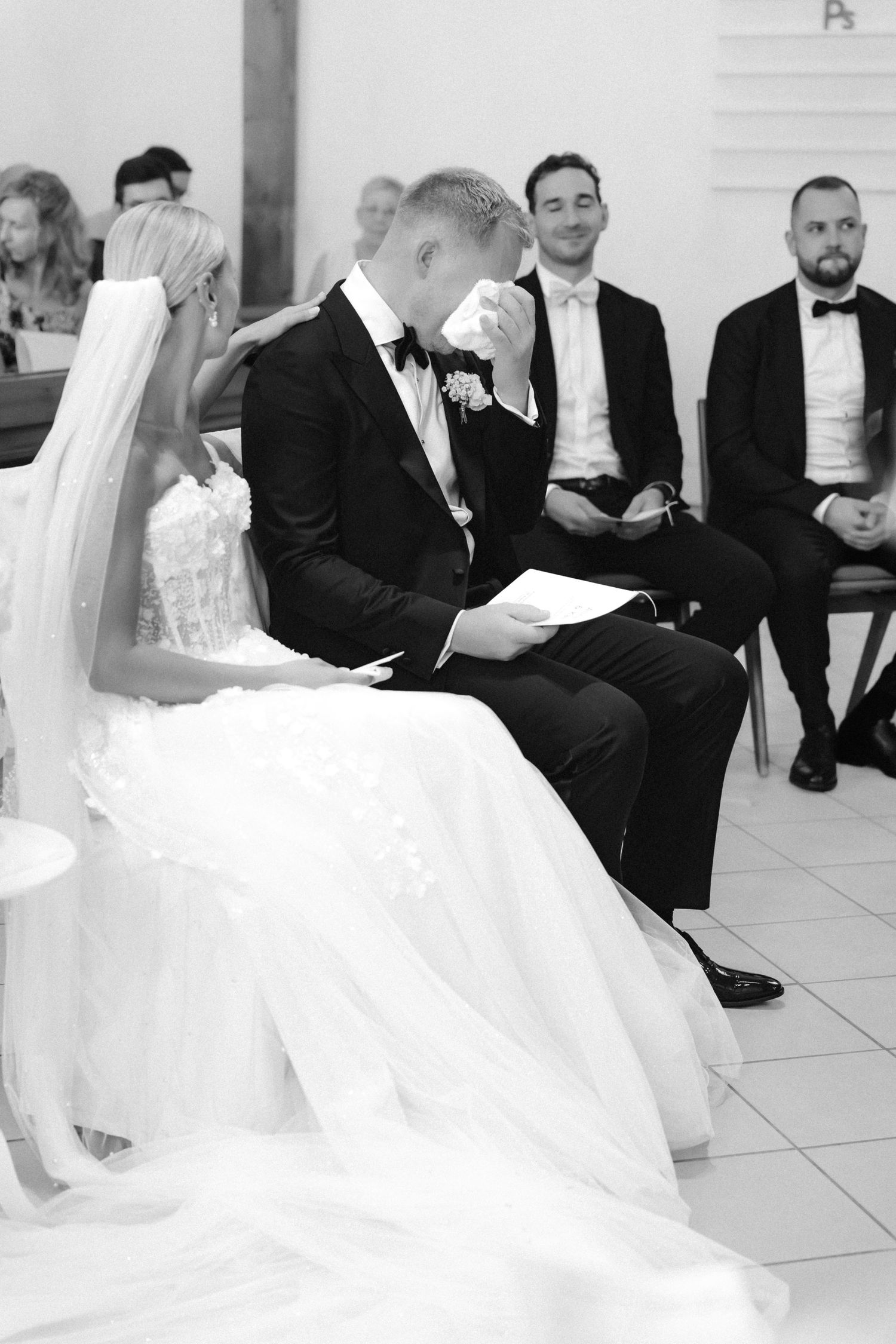 Groom in a black tuxedo wipes tears with a tissue beside the bride in a lace gown and veil during an emotional wedding ceremony.