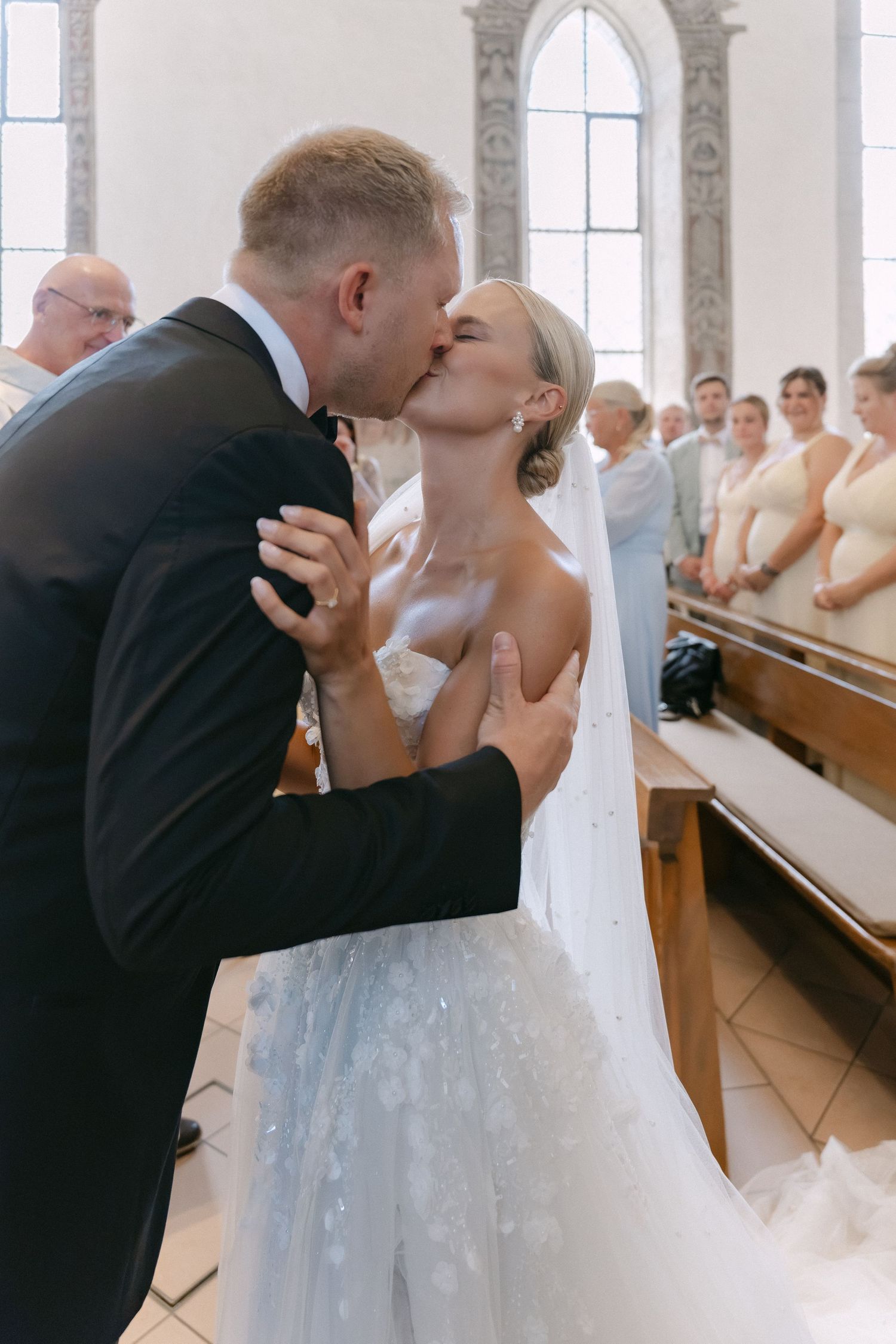 Bride in a lace gown and groom in a dark suit share a wedding kiss in a bright church as guests watch from the pews.