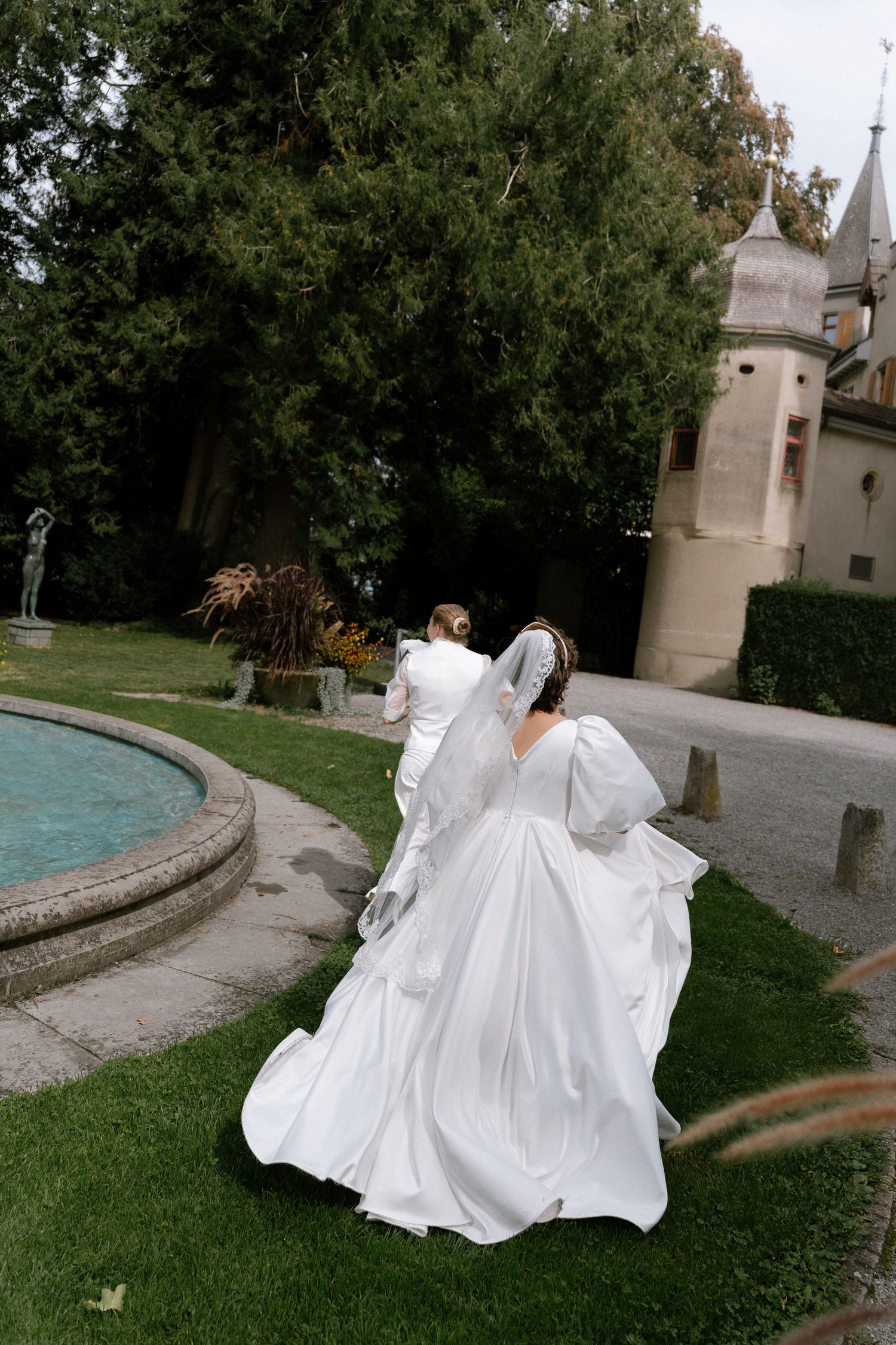 Bride in white gown with veil walks toward castle fountain garden on wedding day.