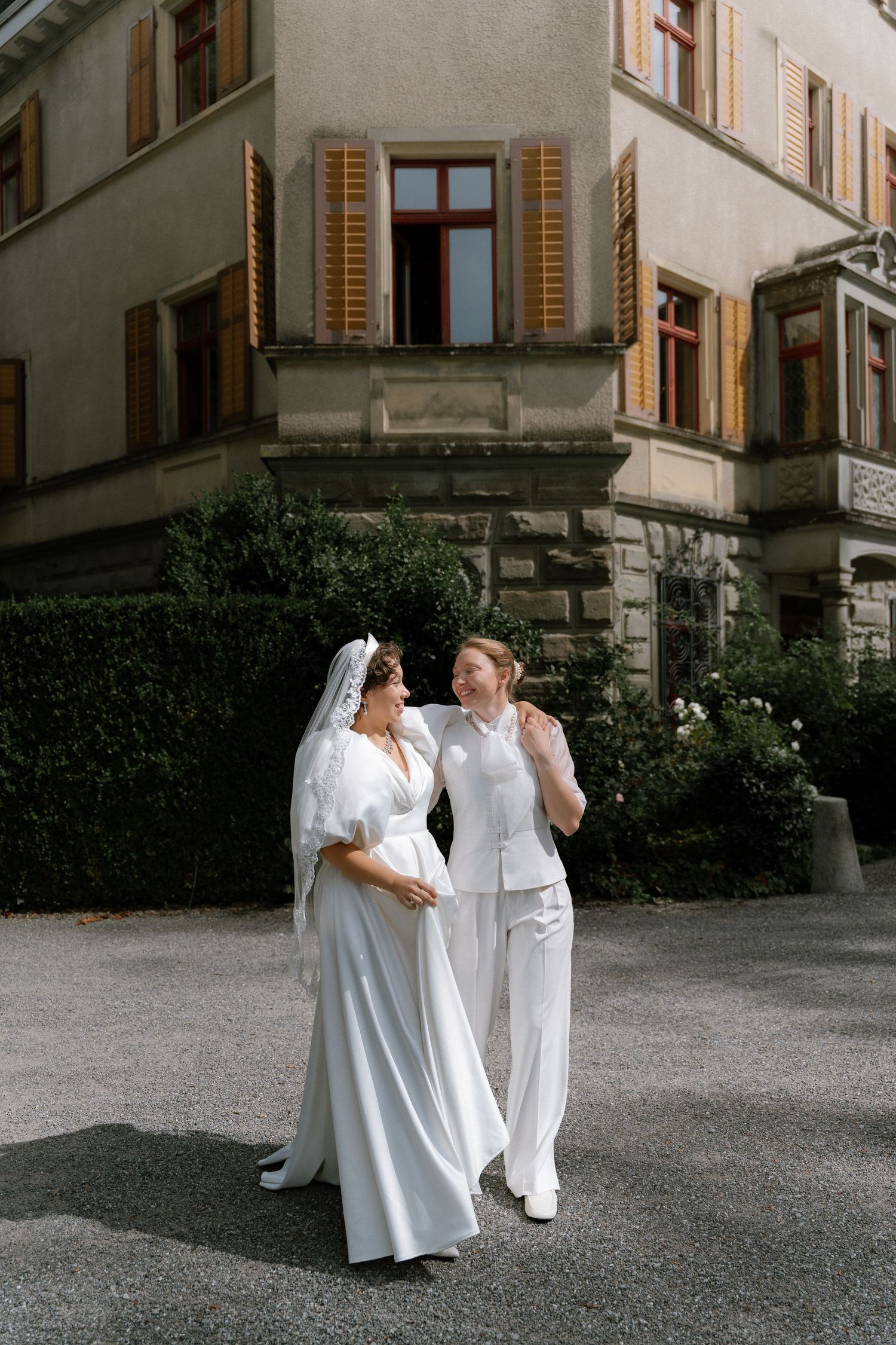 Two brides in white dresses walk together outside a historic building on their wedding day.