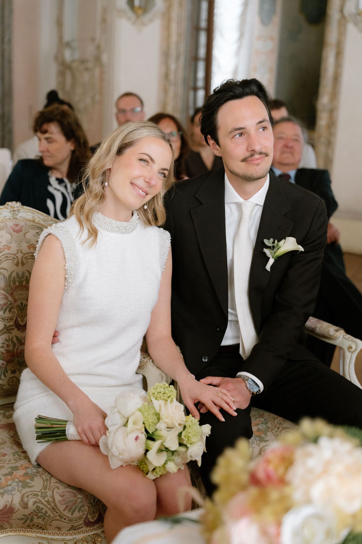 A smiling bride in white dress holds bouquet beside groom in black suit at elegant wedding ceremony.