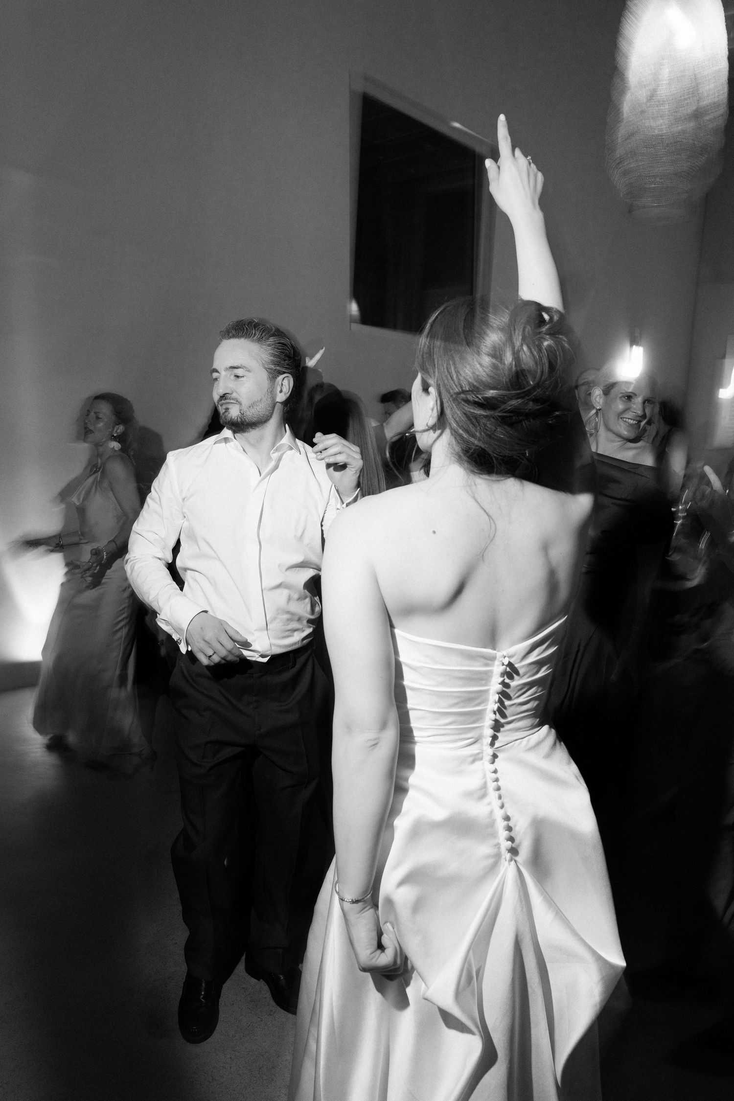 Black and white photo of a bride and groom dancing joyfully at their wedding reception with guests in the background.