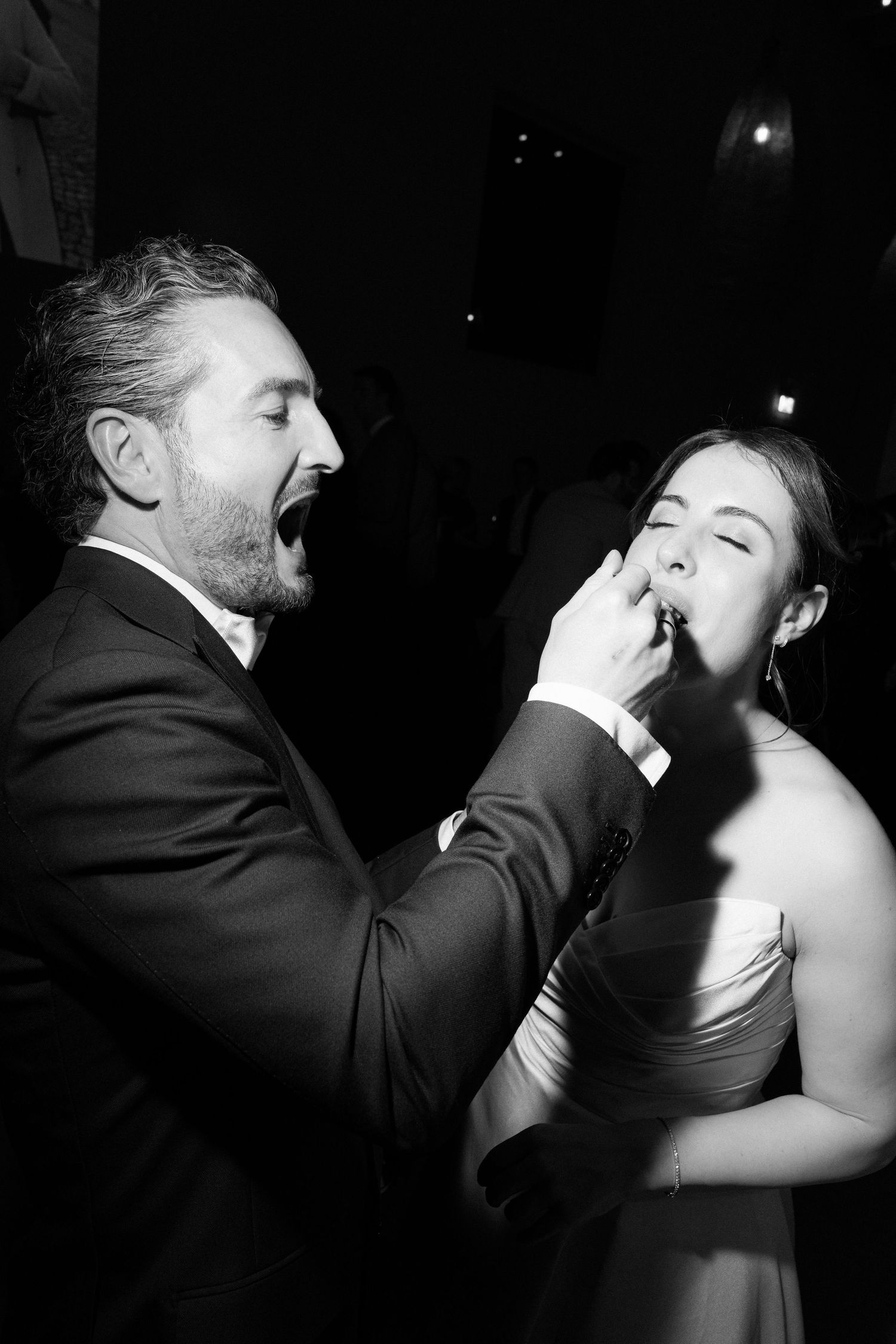 Black and white photo of a man in a suit feeding a woman in a strapless gown at a formal event.