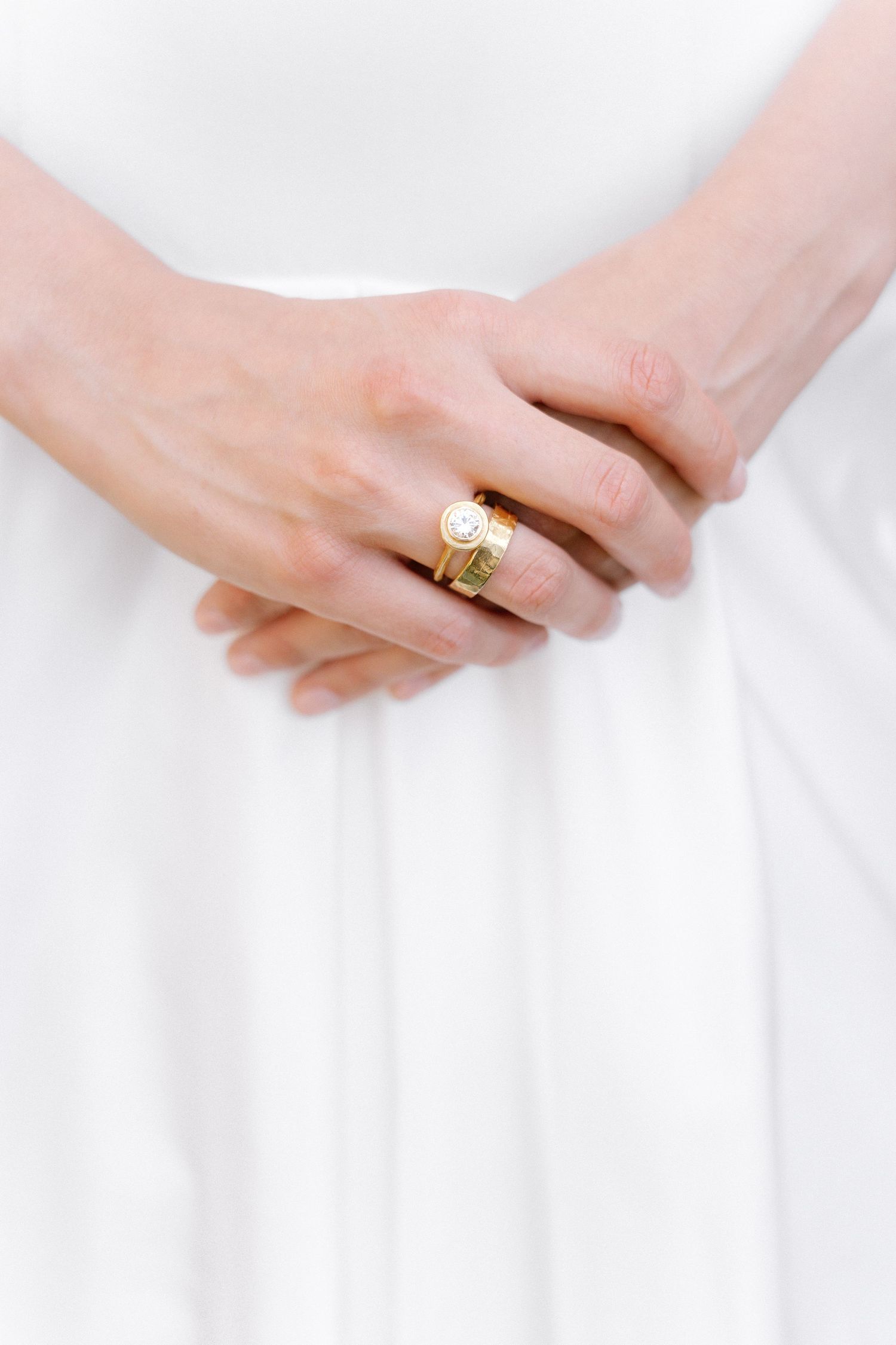 Close-up of bride's hands clasped together wearing stacked gold rings against a white wedding dress.