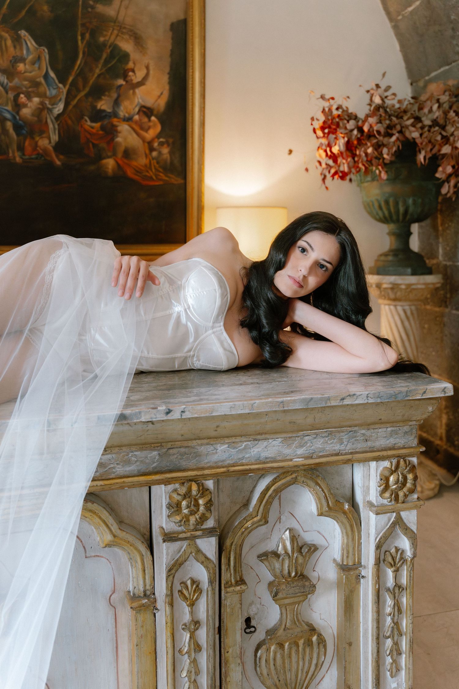 Elegant bride in white tulle gown resting on ornate antique cabinet with classical painting and floral arrangement.
