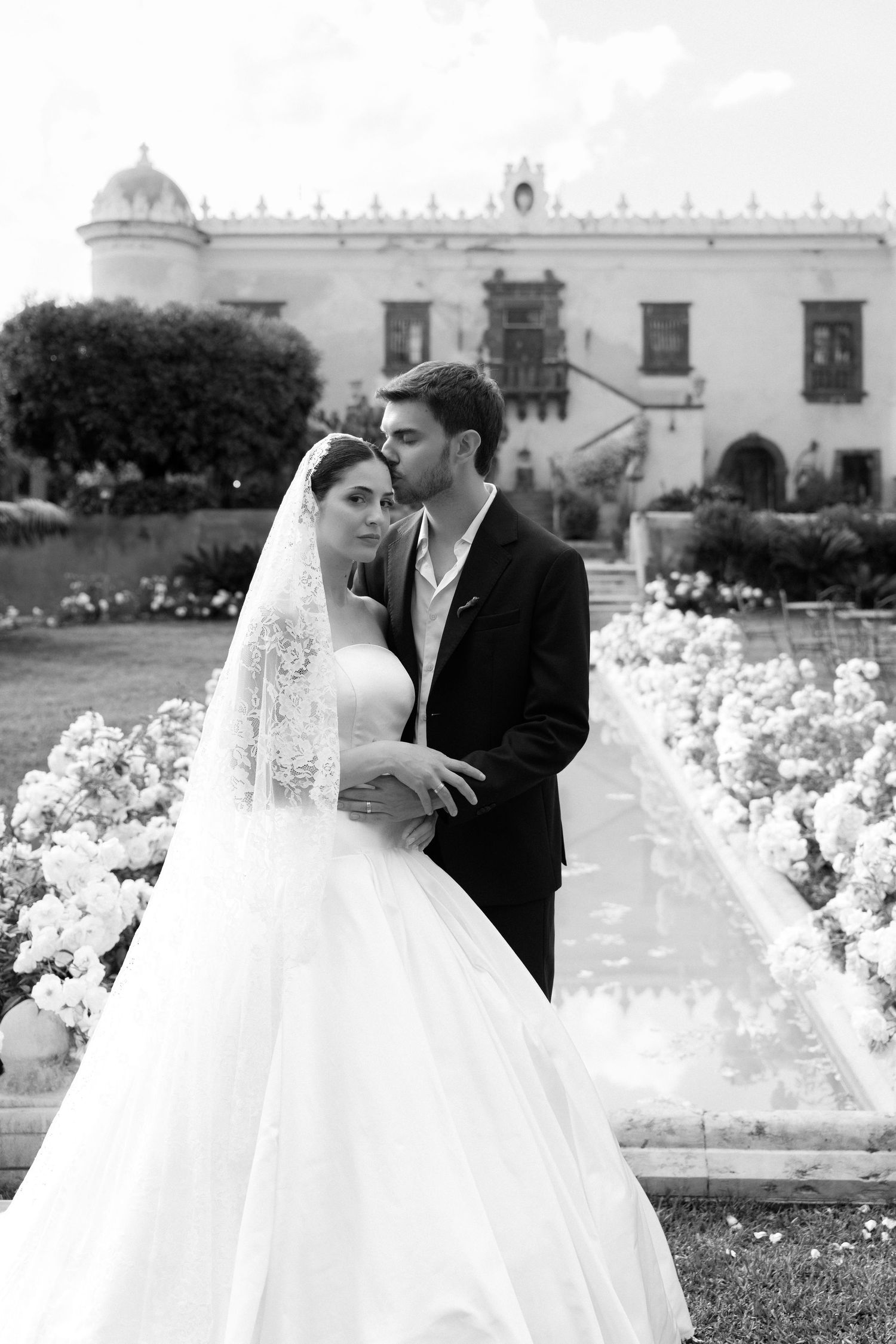 Black and white wedding photo of bride in ball gown and groom in suit posing before an elegant estate garden.