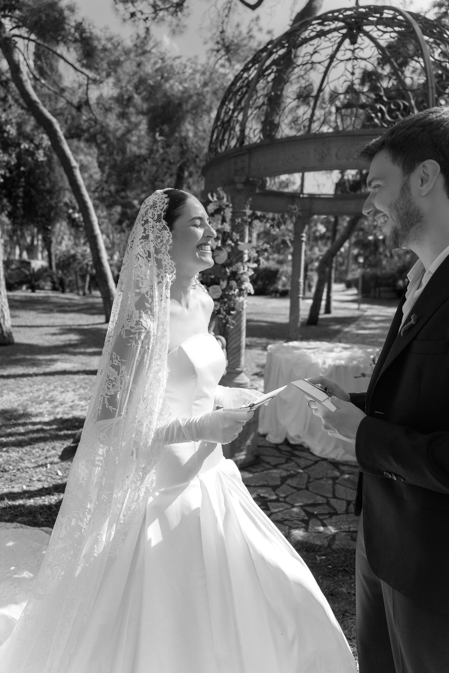 Black and white photo of a smiling bride in a lace veil reading vows to her groom in a sunlit garden.