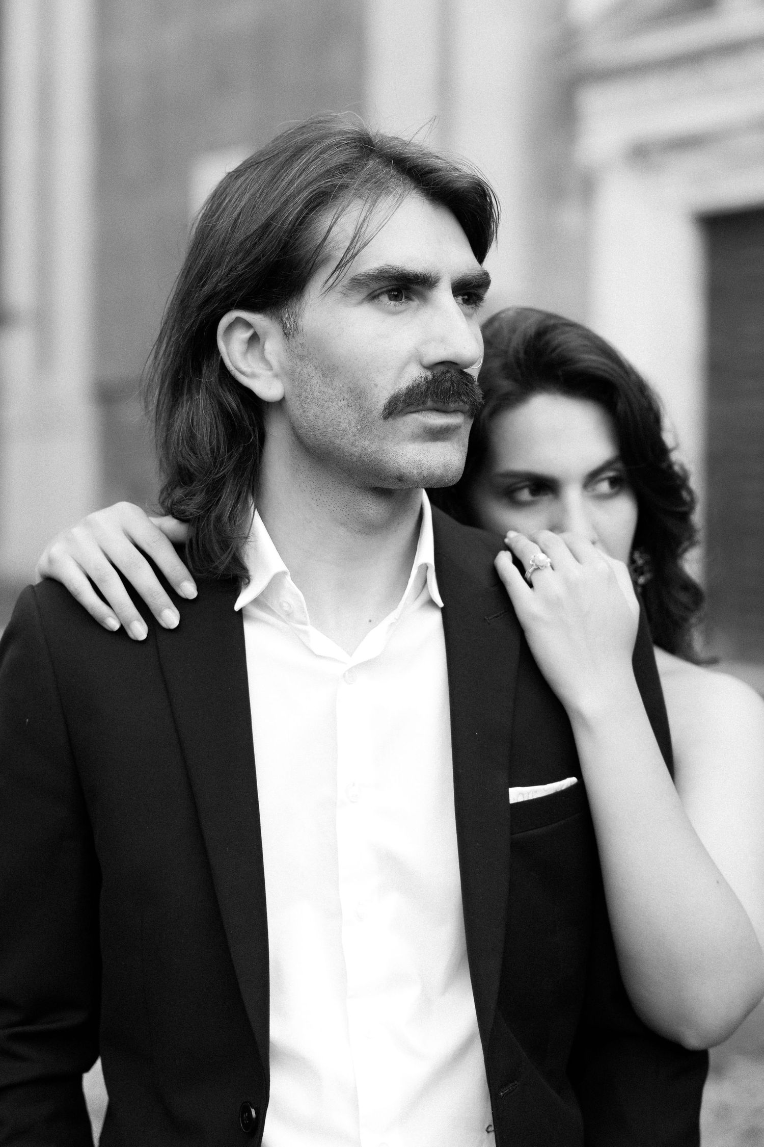 Black and white portrait of elegant couple, man with mustache in suit jacket, woman resting hand on his shoulder.