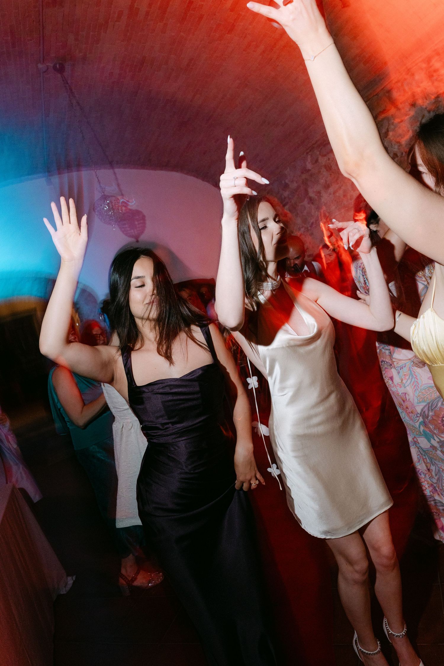 Women in elegant dresses dancing energetically at a party under colorful red and blue lights.