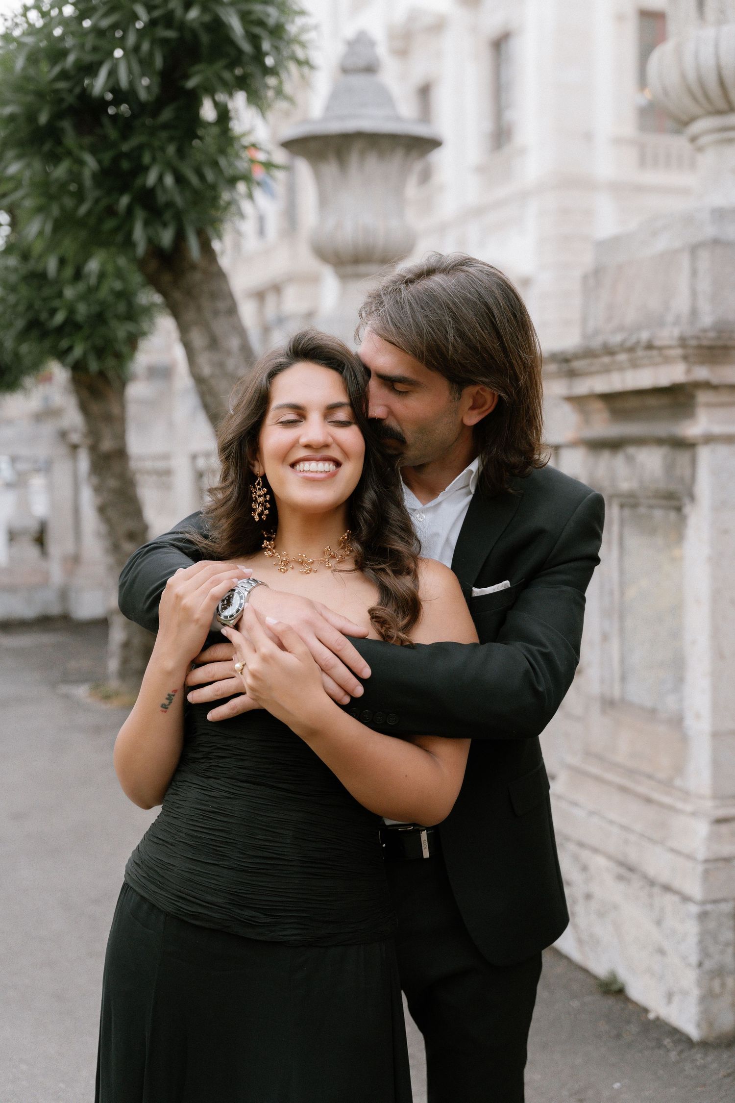 A couple in elegant black attire embraces outdoors, the man kissing the woman's head as she smiles joyfully.