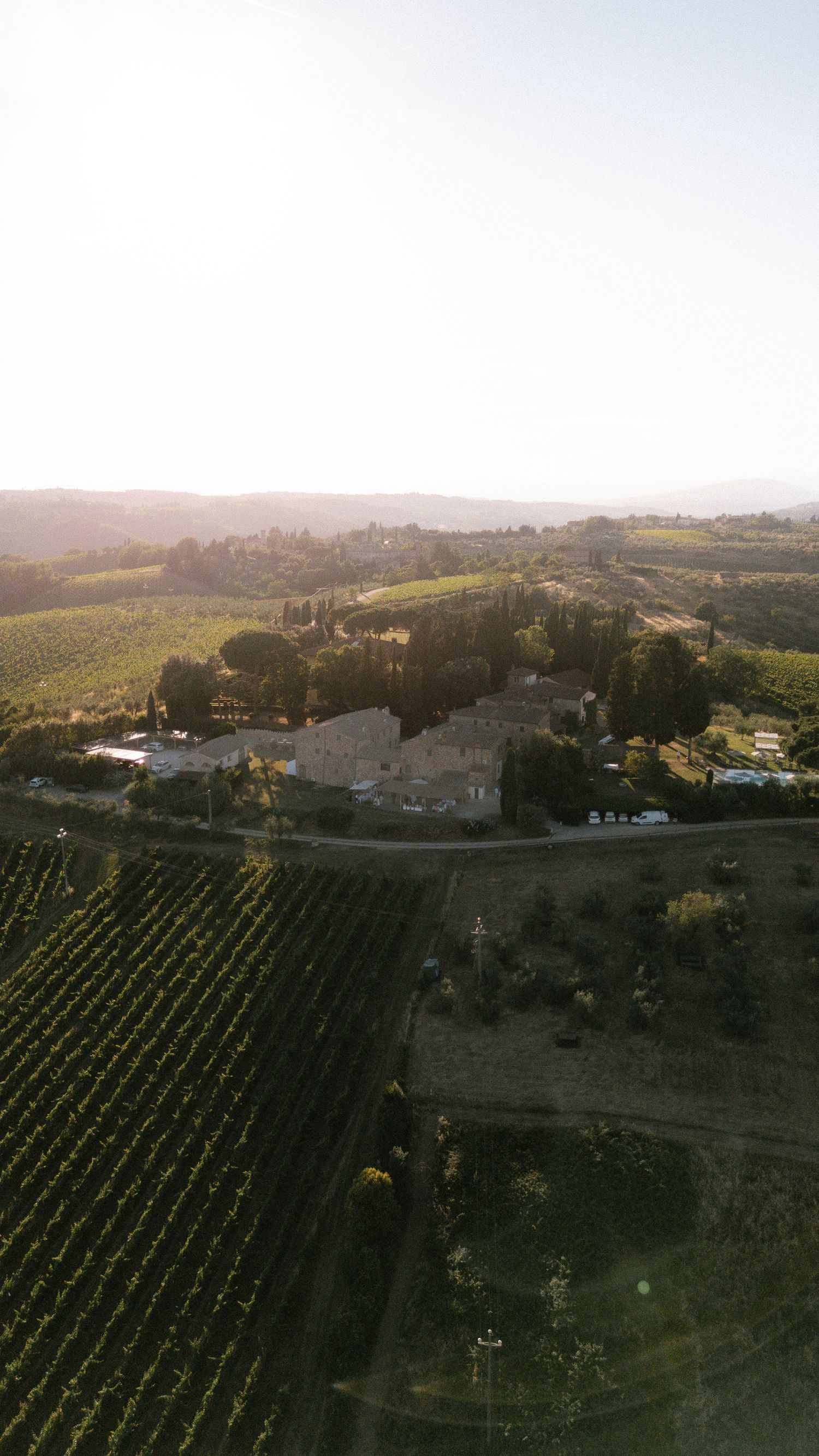 Aerial drone view of a Tuscan vineyard estate with rolling green hills, cypress trees, and a stone farmhouse at golden hour.