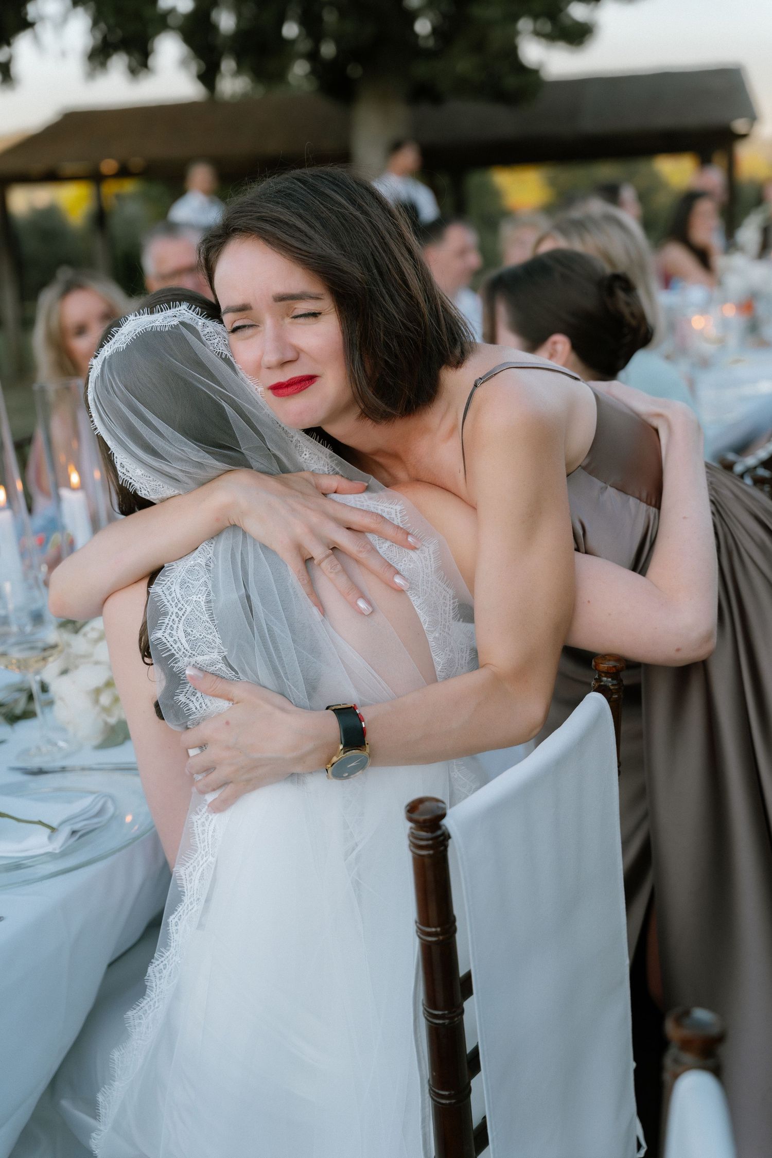 A woman in a gray dress emotionally hugs a bride in a white gown and veil at an outdoor wedding reception.