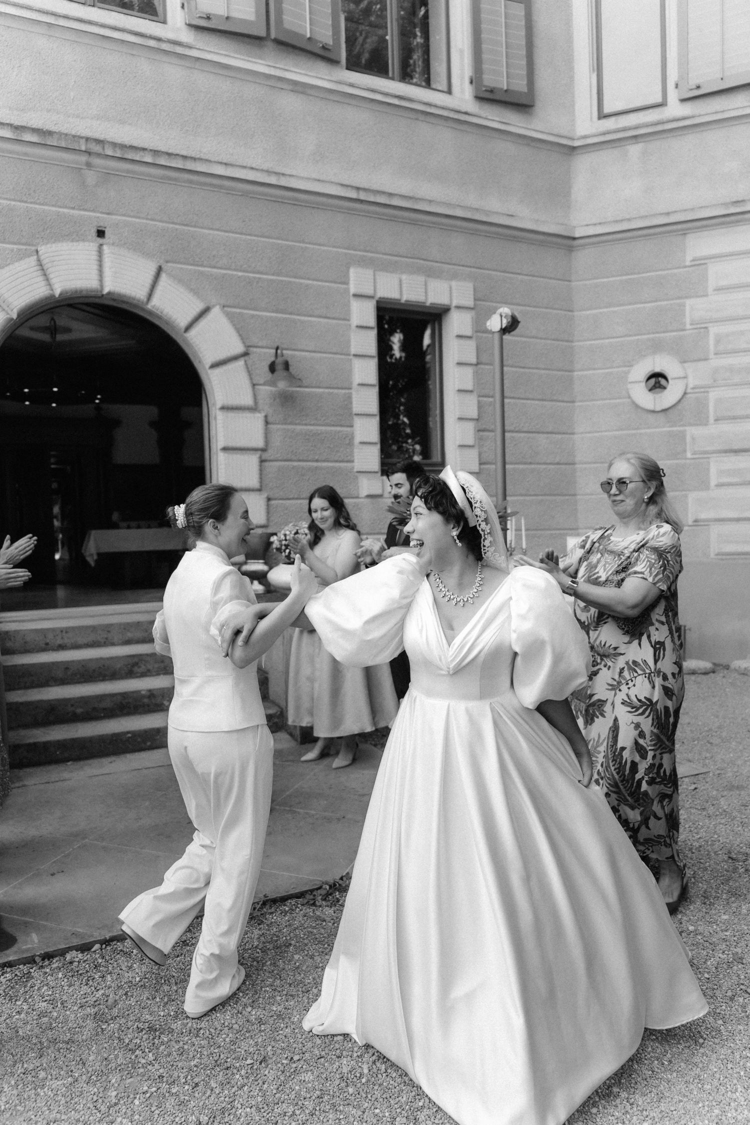 Black and white photo of a bride in a white gown dancing with guests outside a elegant stone building.