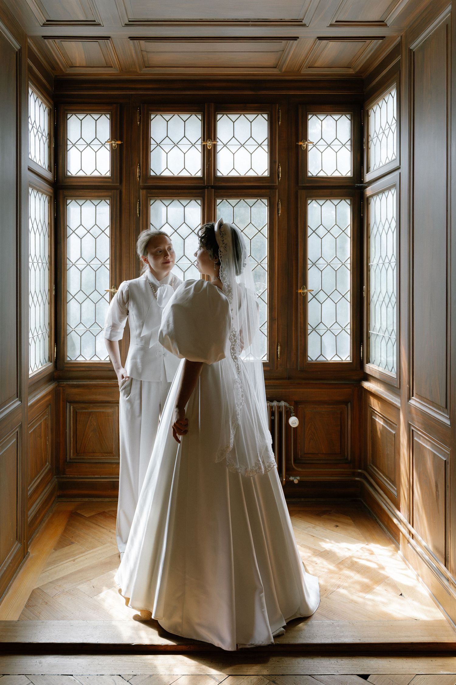 Bride and groom stand facing each other in elegant wood-paneled room with leaded glass windows, bathed in soft natural light.