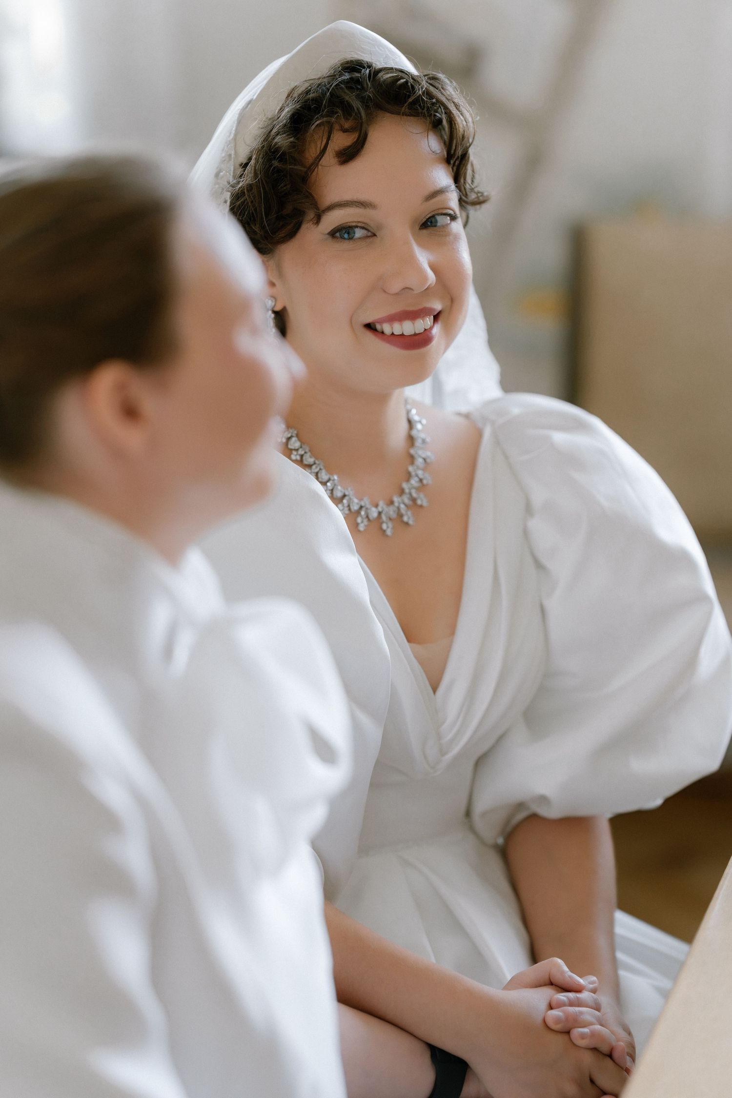 Bride in white gown with puff sleeves, diamond necklace, and veil smiles warmly at another woman in white.