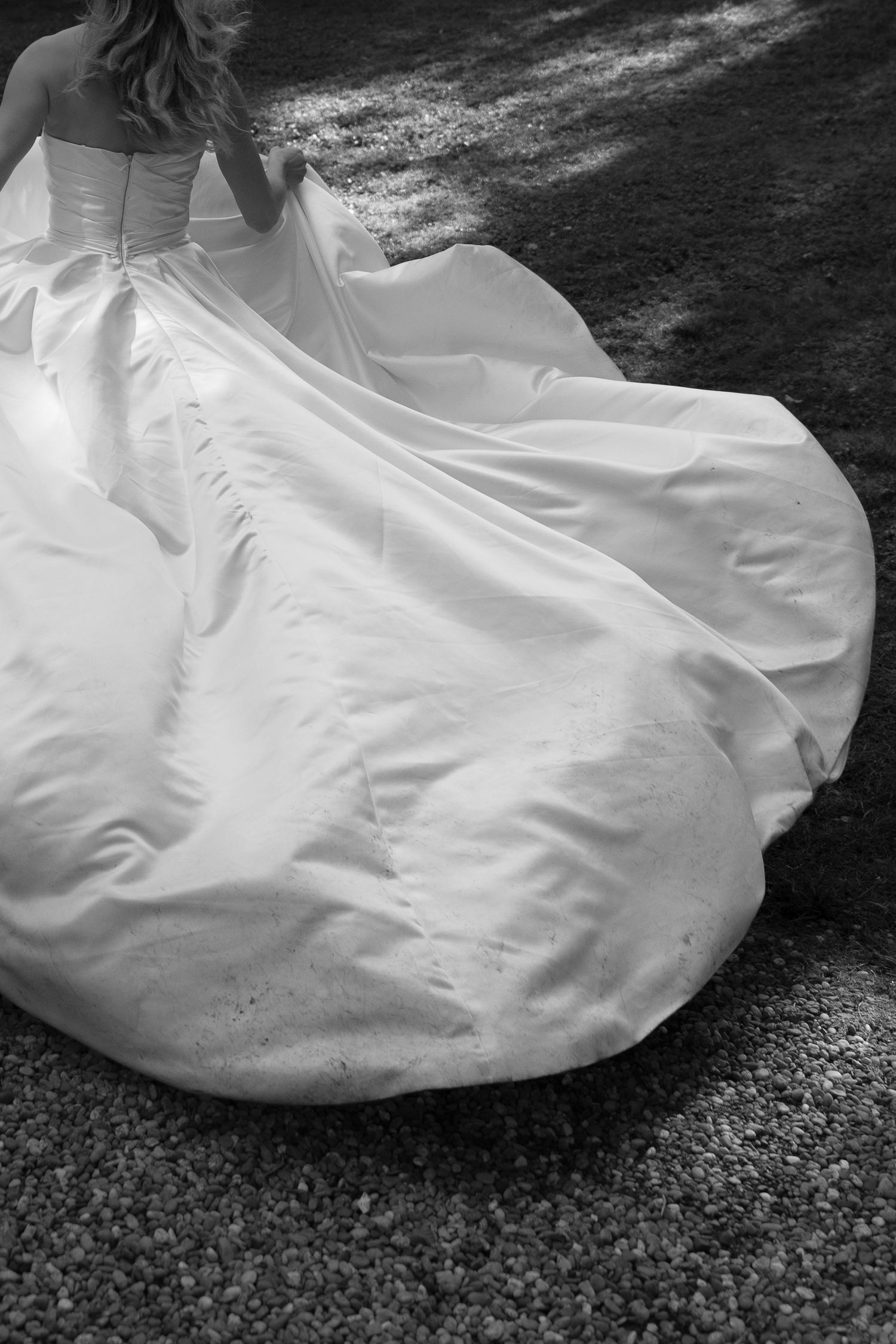 Black and white photo of a bride in a strapless satin wedding gown with a full flowing skirt, walking across a gravel path.