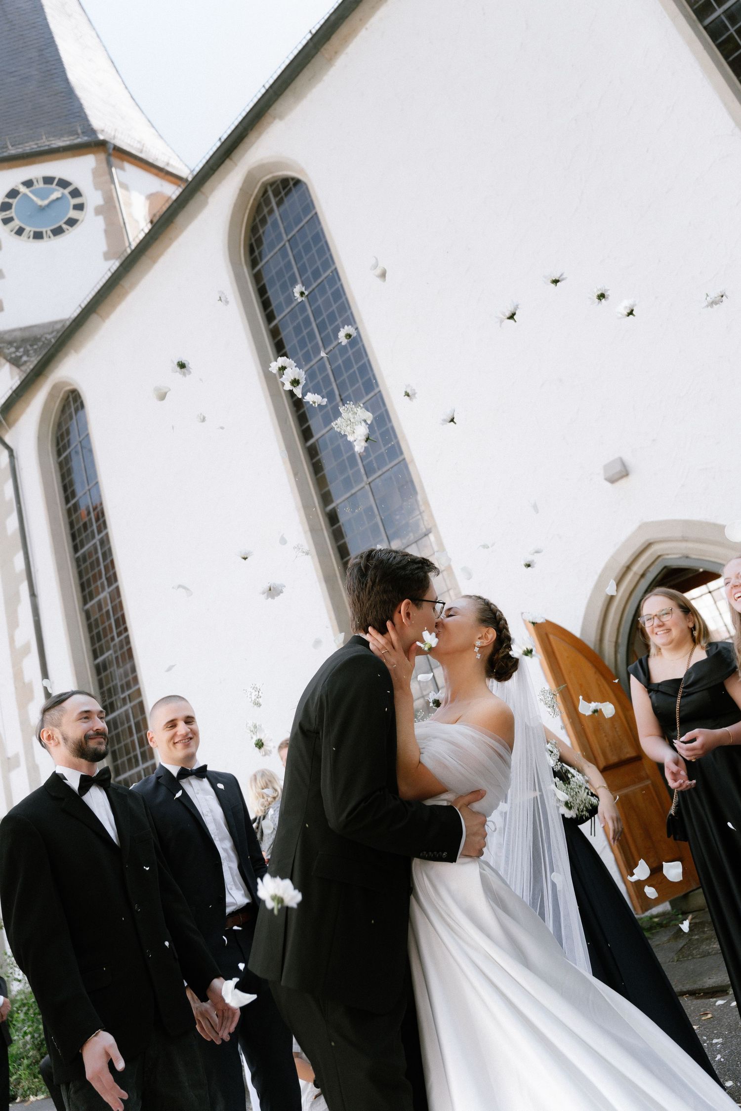 Bride and groom share a kiss outside a white church as groomsmen and wedding guests look on, with confetti falling around them.
