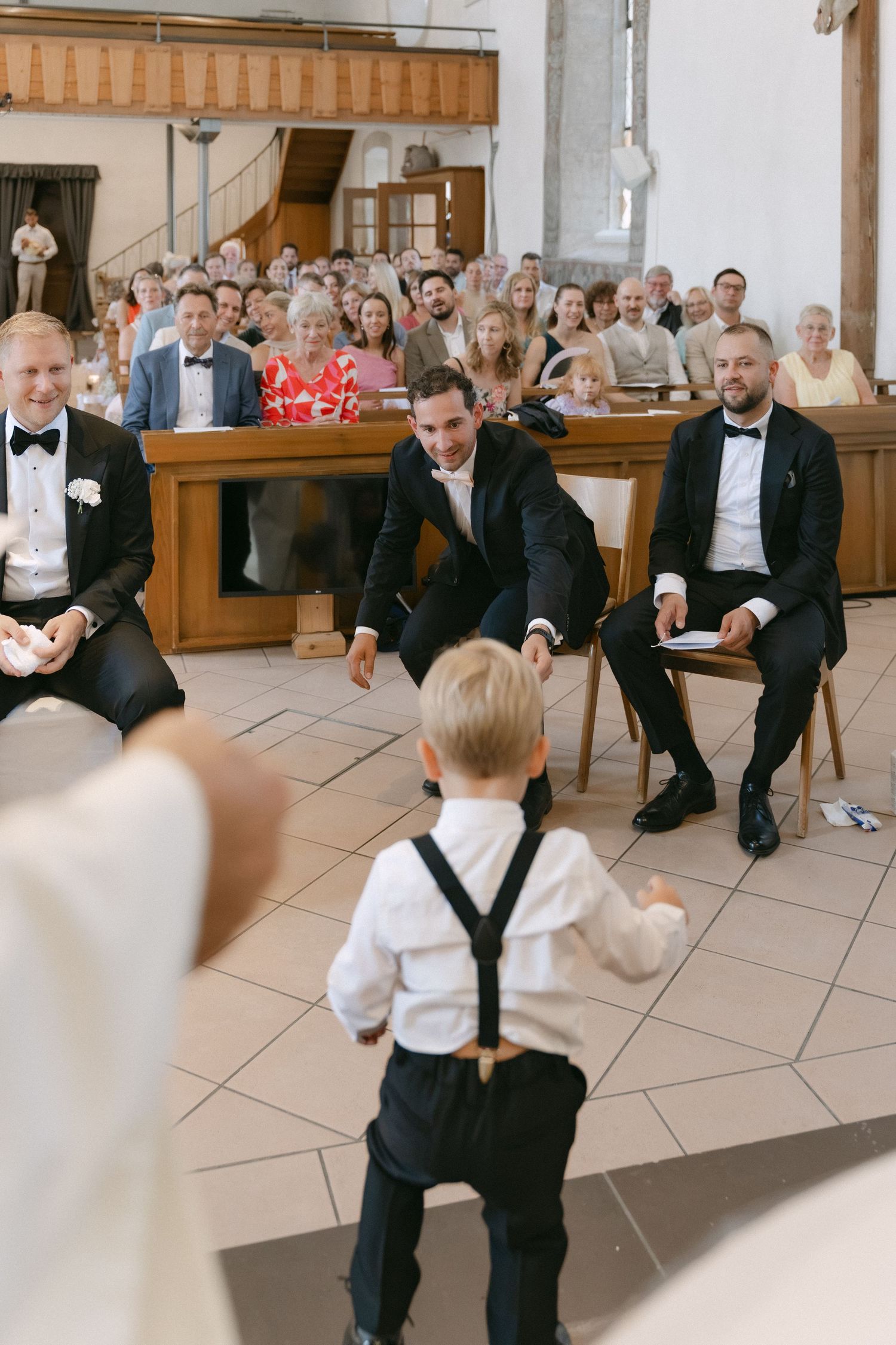 Little boy in suspenders walks across a church floor toward smiling groomsmen as wedding guests watch from the pews.