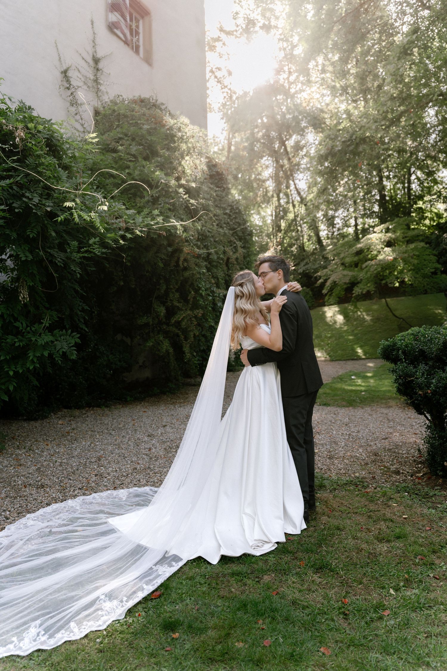Bride in a white gown with a long cathedral veil embraces her groom in a black suit in a sunlit garden surrounded by lush greenery.