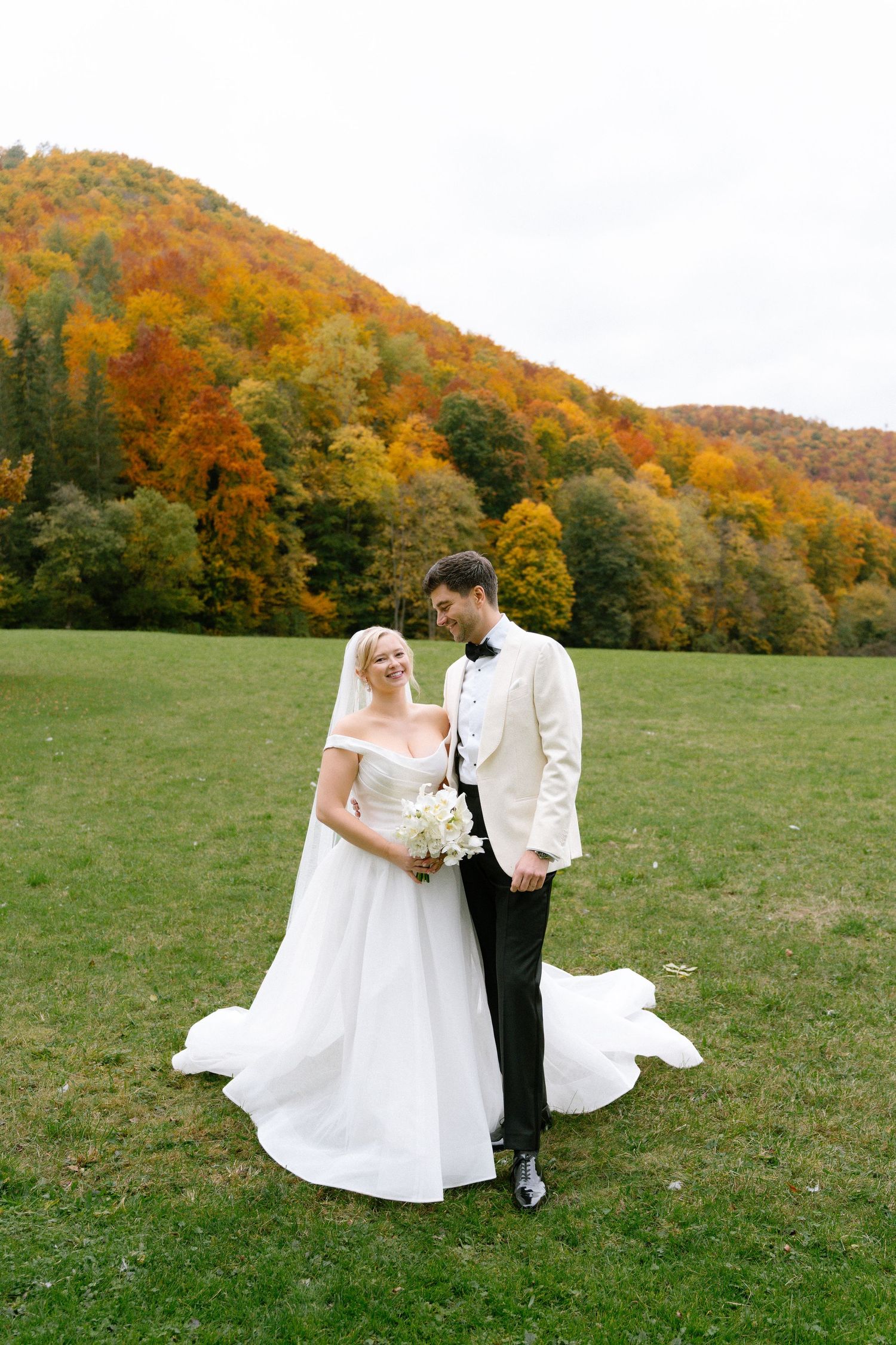 A couple in wedding attire stands on a grassy field with vibrant autumn foliage on rolling hills in the background.